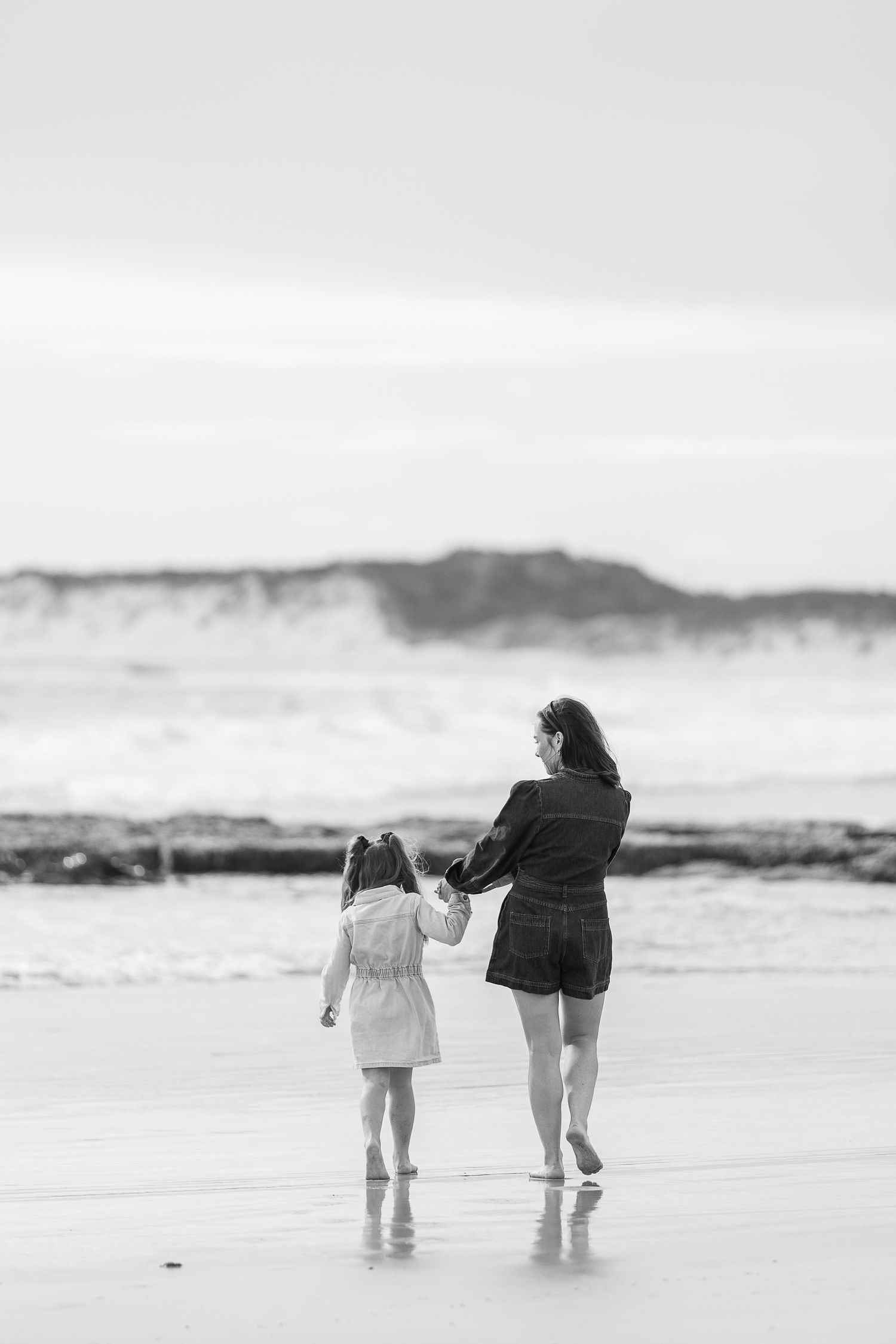 A parent and child walk hand in hand along the beach shore in black and white.