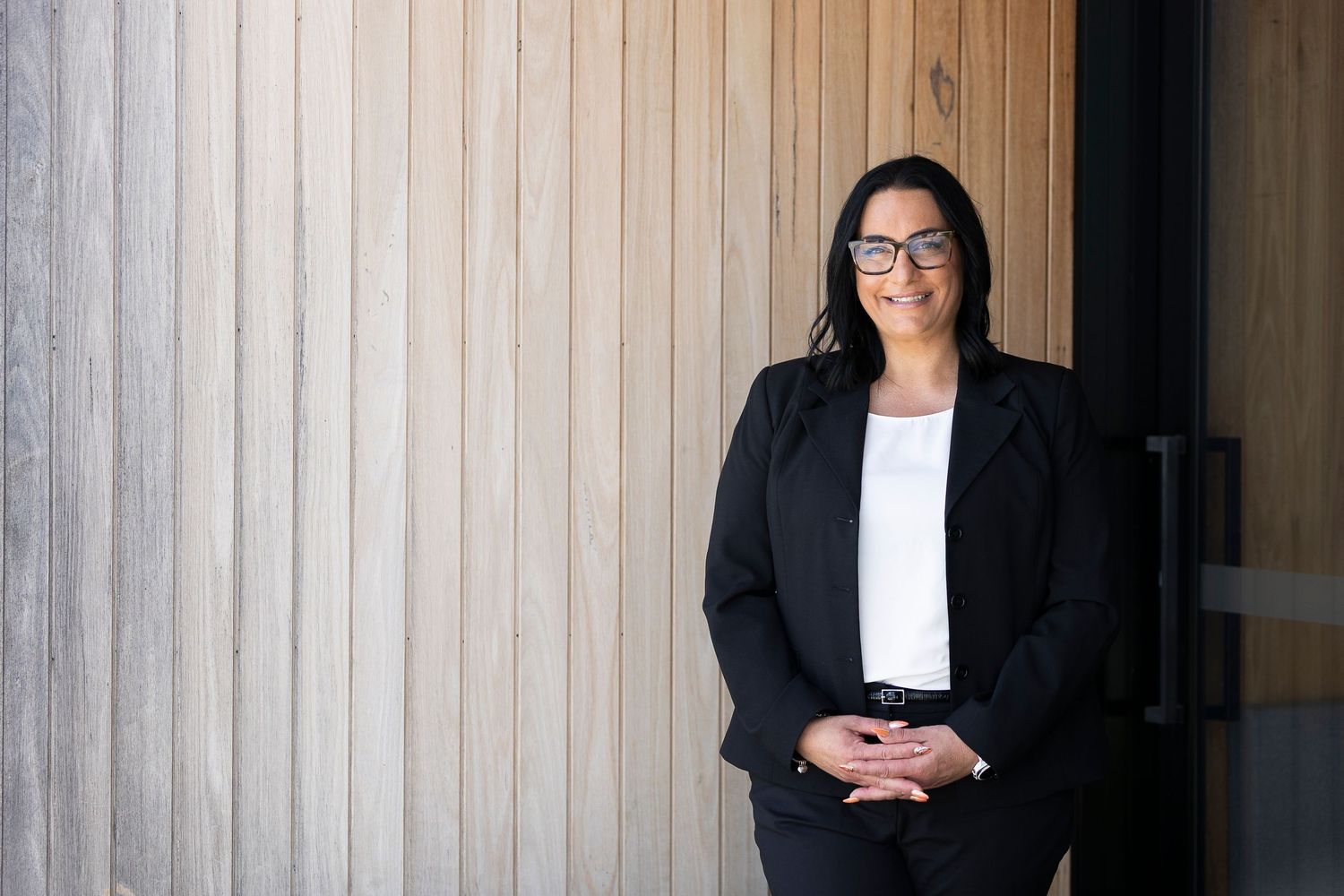 Professional wearing glasses and black blazer seated against wooden wall.