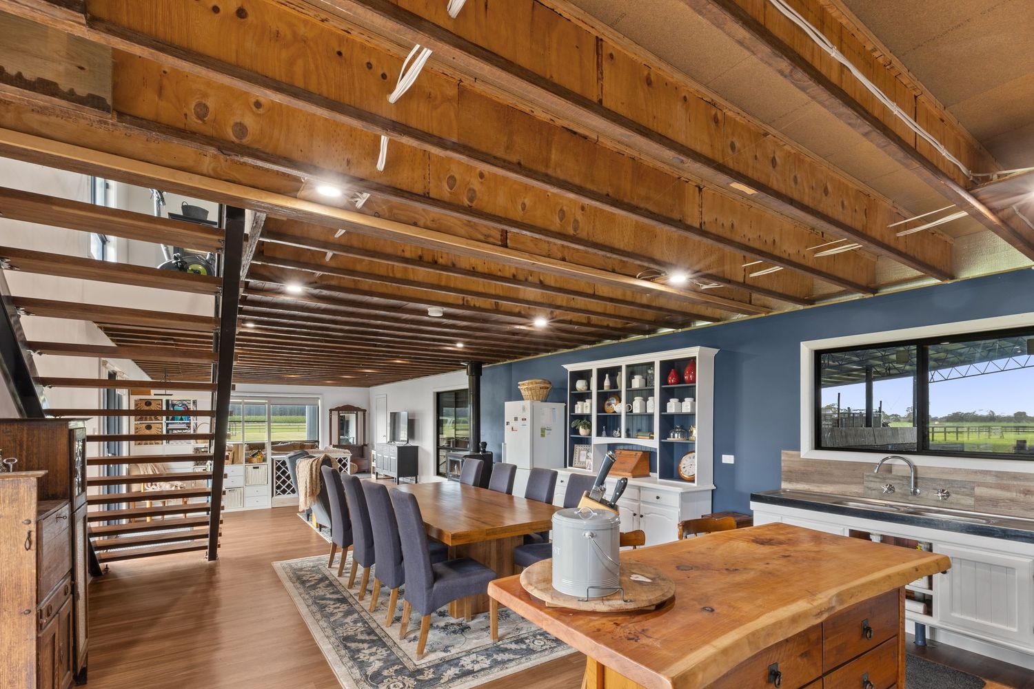 Rustic open-plan dining area with wooden ceiling beams, large farmhouse table, blue accent wall, and modern kitchen with natural light.