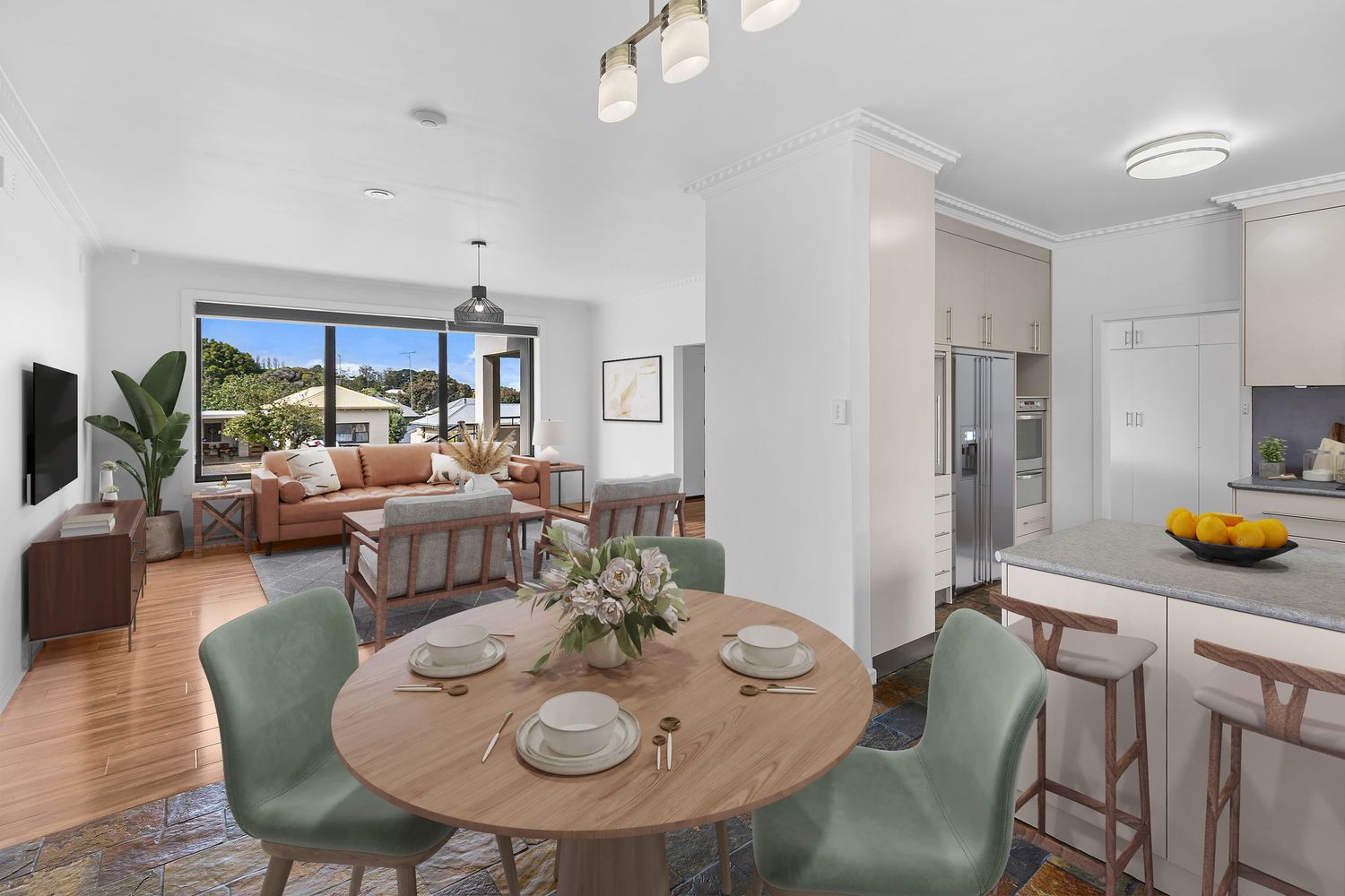 Modern open-plan dining and living room with round wooden table, green chairs, and kitchen with stainless steel appliances.