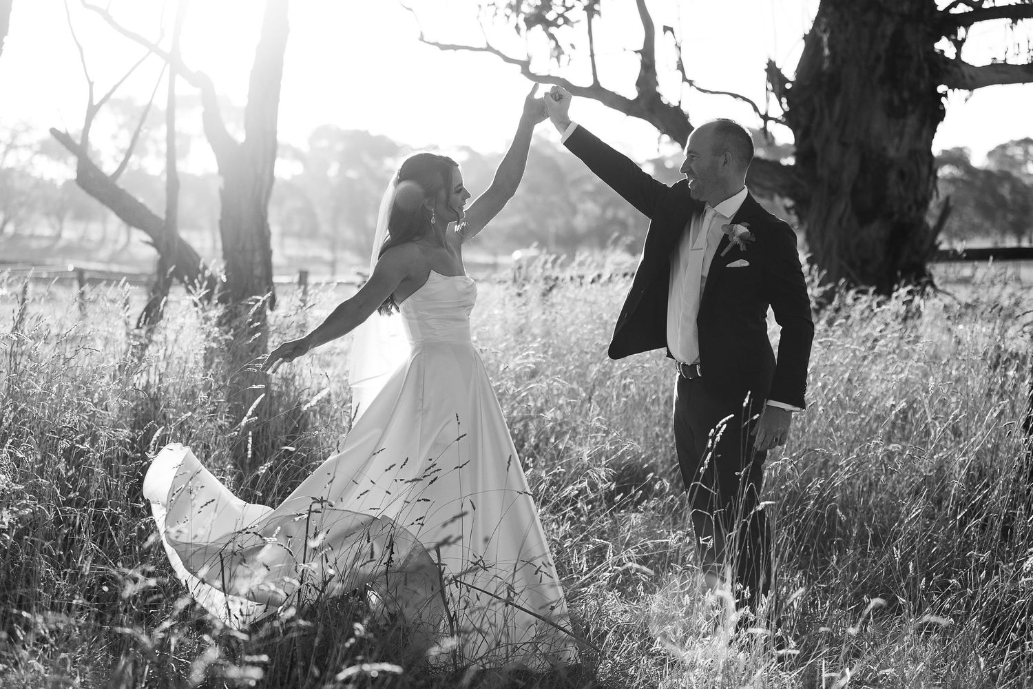 A joyful couple dances together in a sunlit field with tall grass and trees.