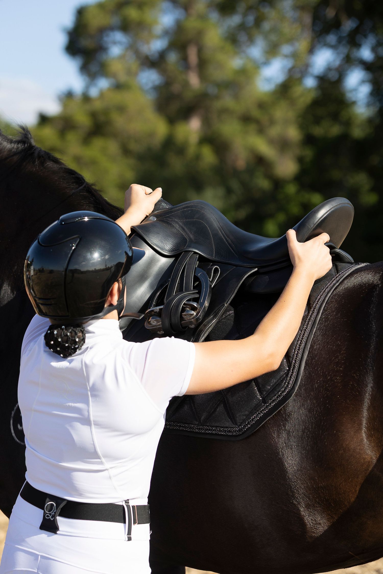 Close-up of equestrian adjusting saddle on black horse.