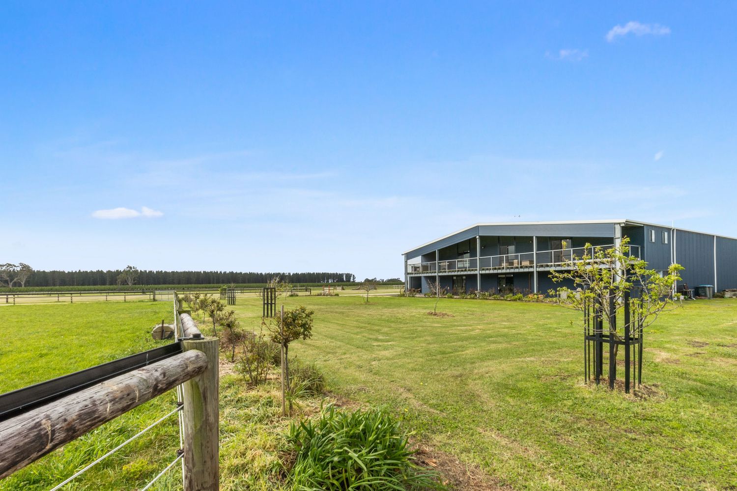 Rural property featuring a modern two-story home with deck, lush green paddocks, wooden fence, and tree-lined horizon under blue sky.