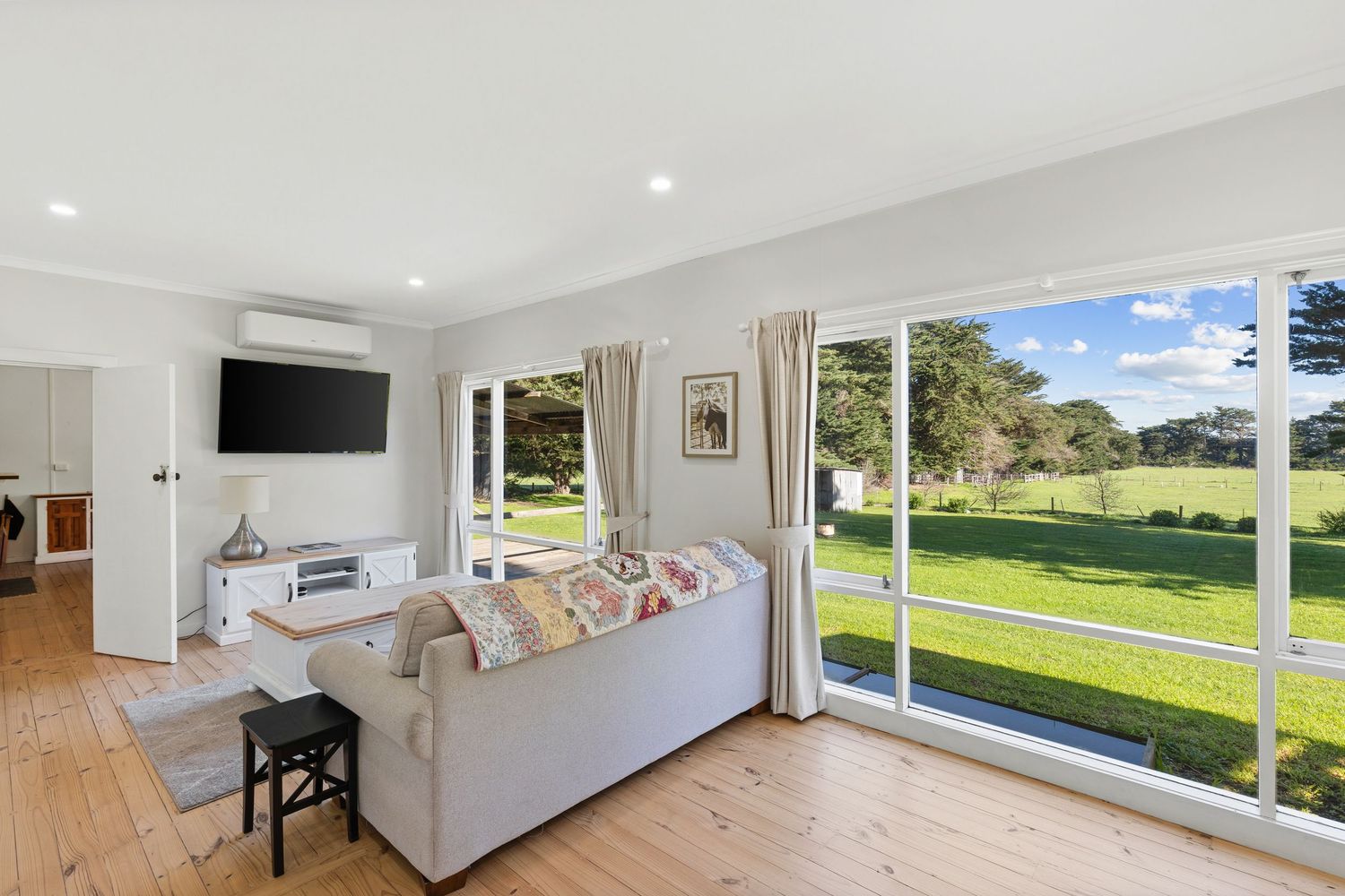 Bright living room with white sofa, wall-mounted TV, hardwood floors, and large sliding doors overlooking a lush green lawn.