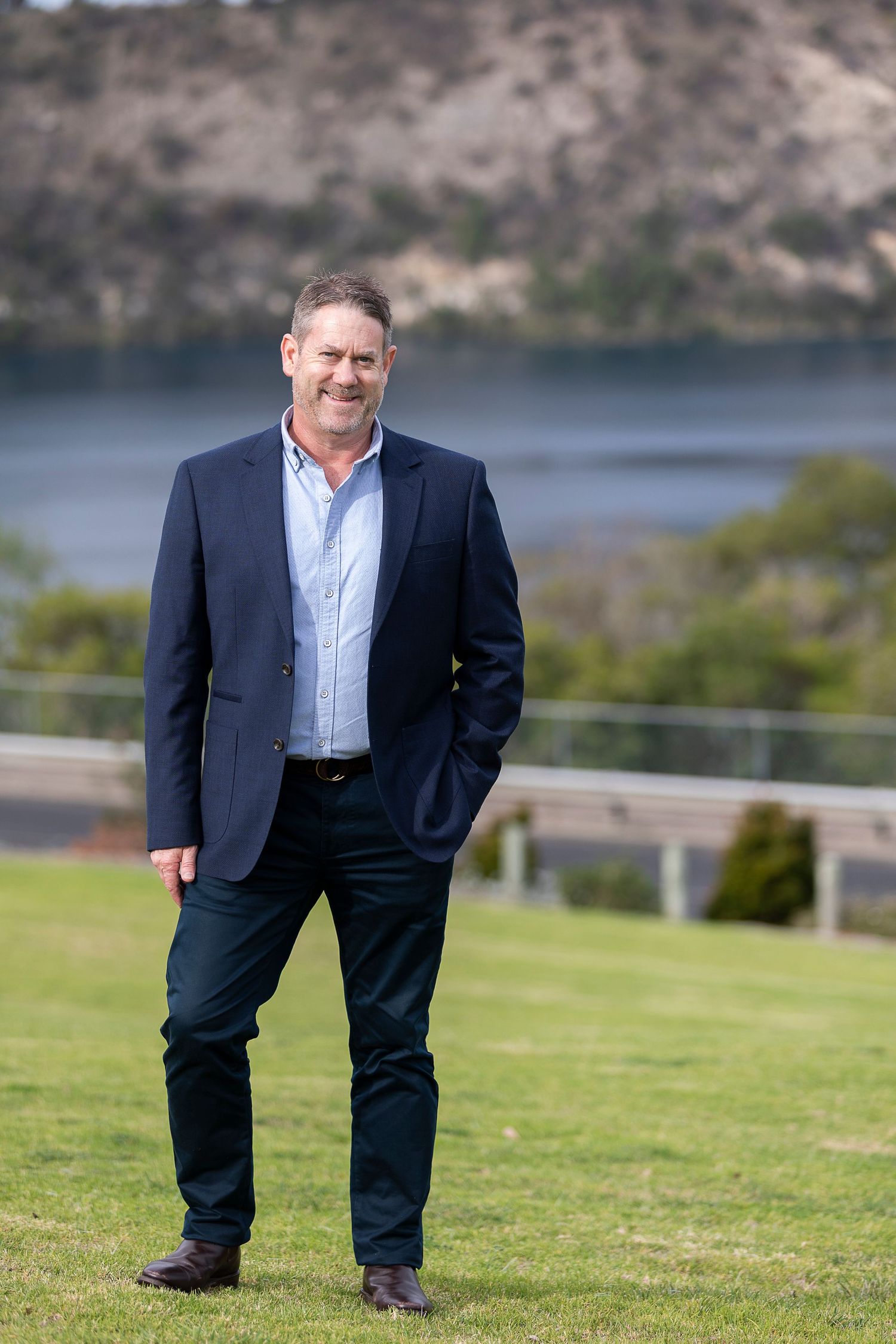 Smiling middle-aged man in navy blazer and dark trousers standing on green lawn with coastal scenery behind him.