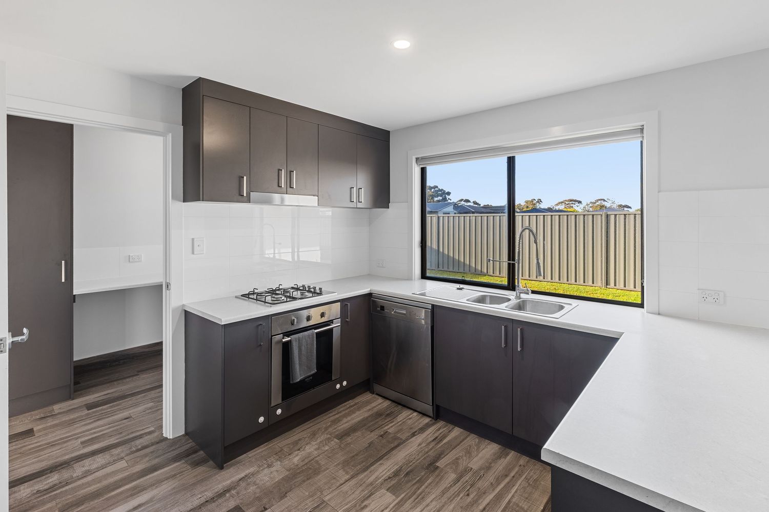 Modern kitchen with dark cabinets, stainless steel appliances, and wood-look flooring extends to living area.