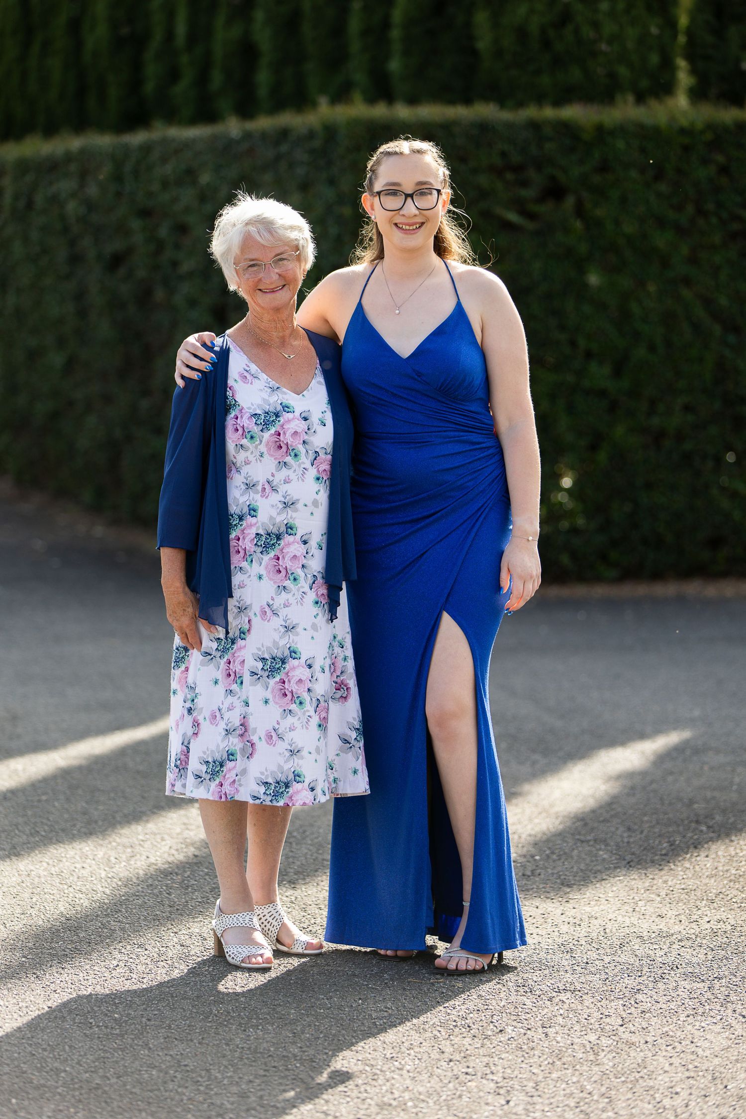 Two people pose together outdoors, one wearing a floral dress and the other in a royal blue formal gown.