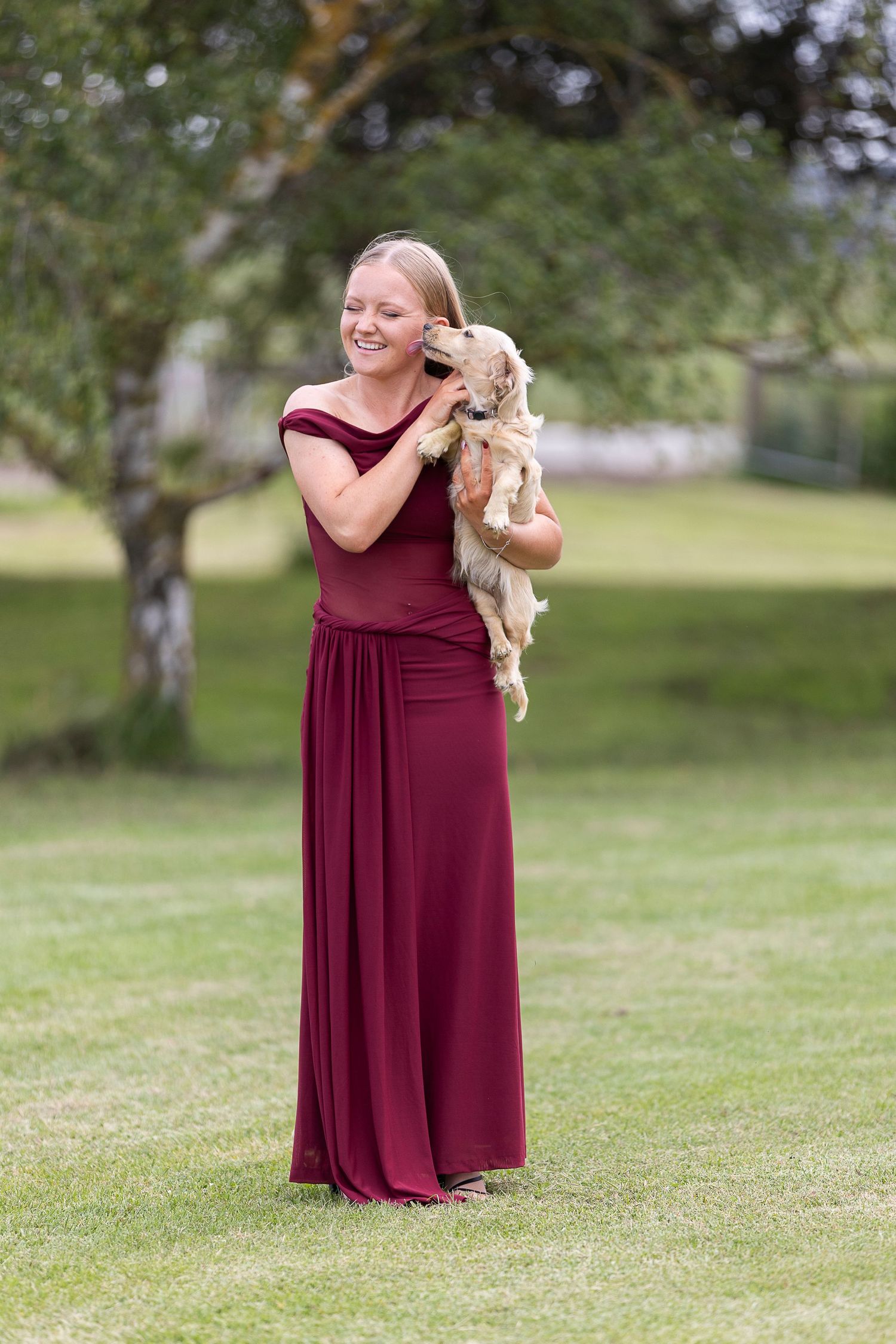 A woman in a burgundy dress holds a small light-colored puppy while standing on grass.