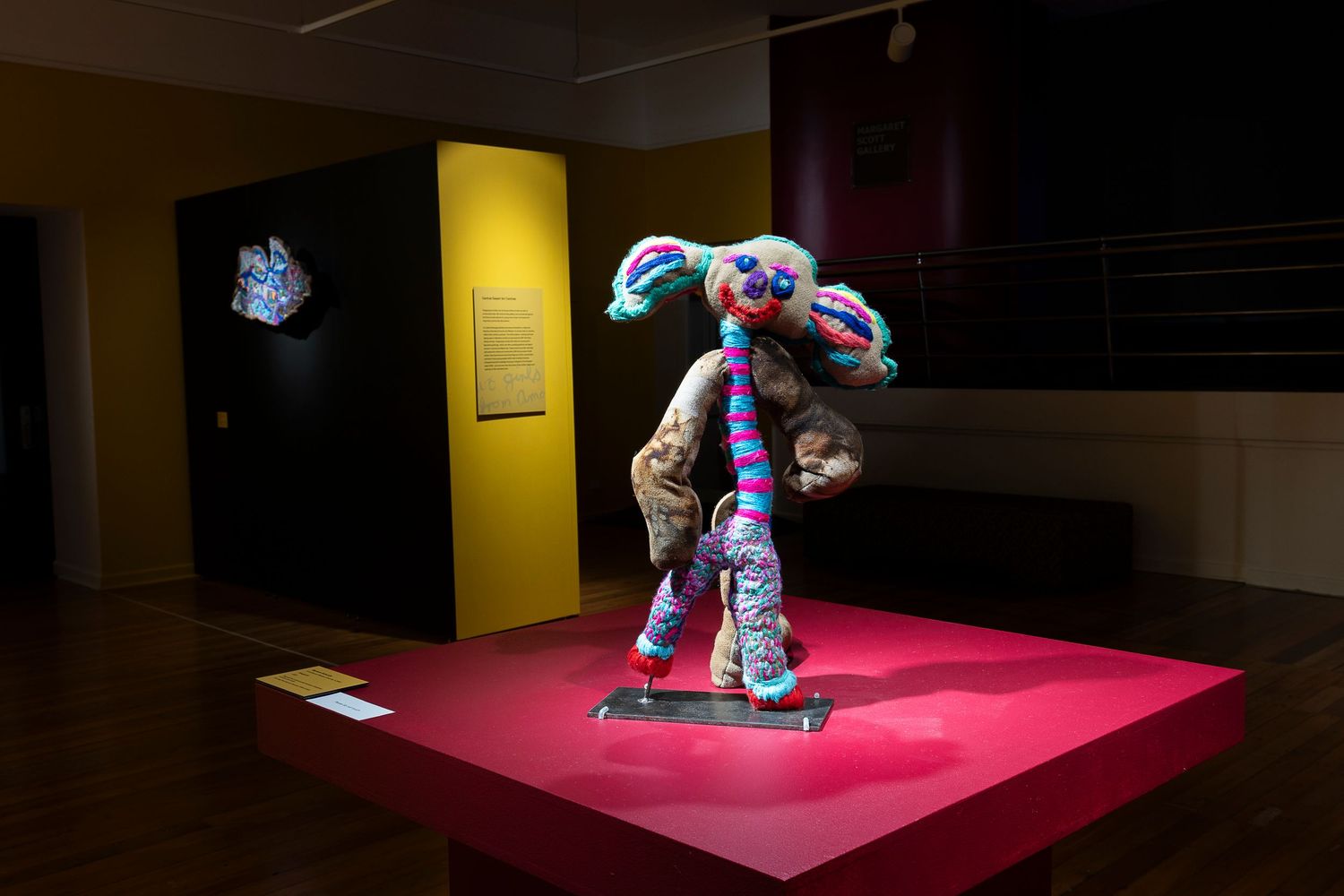 Art gallery exhibition space with pink pedestals displaying sculptural works against yellow walls.