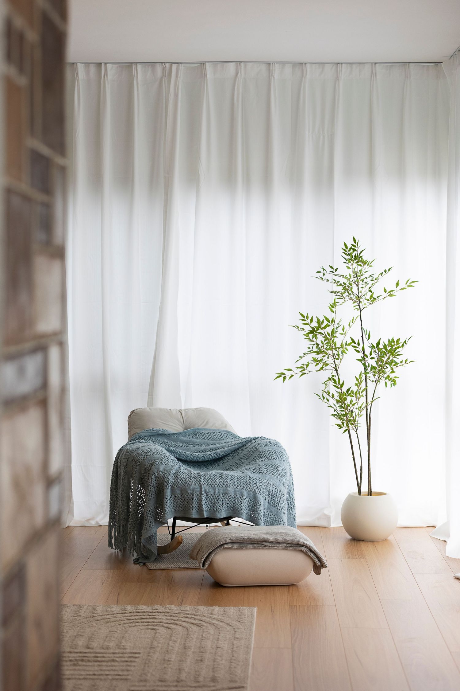 Minimalist living room with teal throw on grey chair, white curtains, wood floor, and tall green plant in white pot.