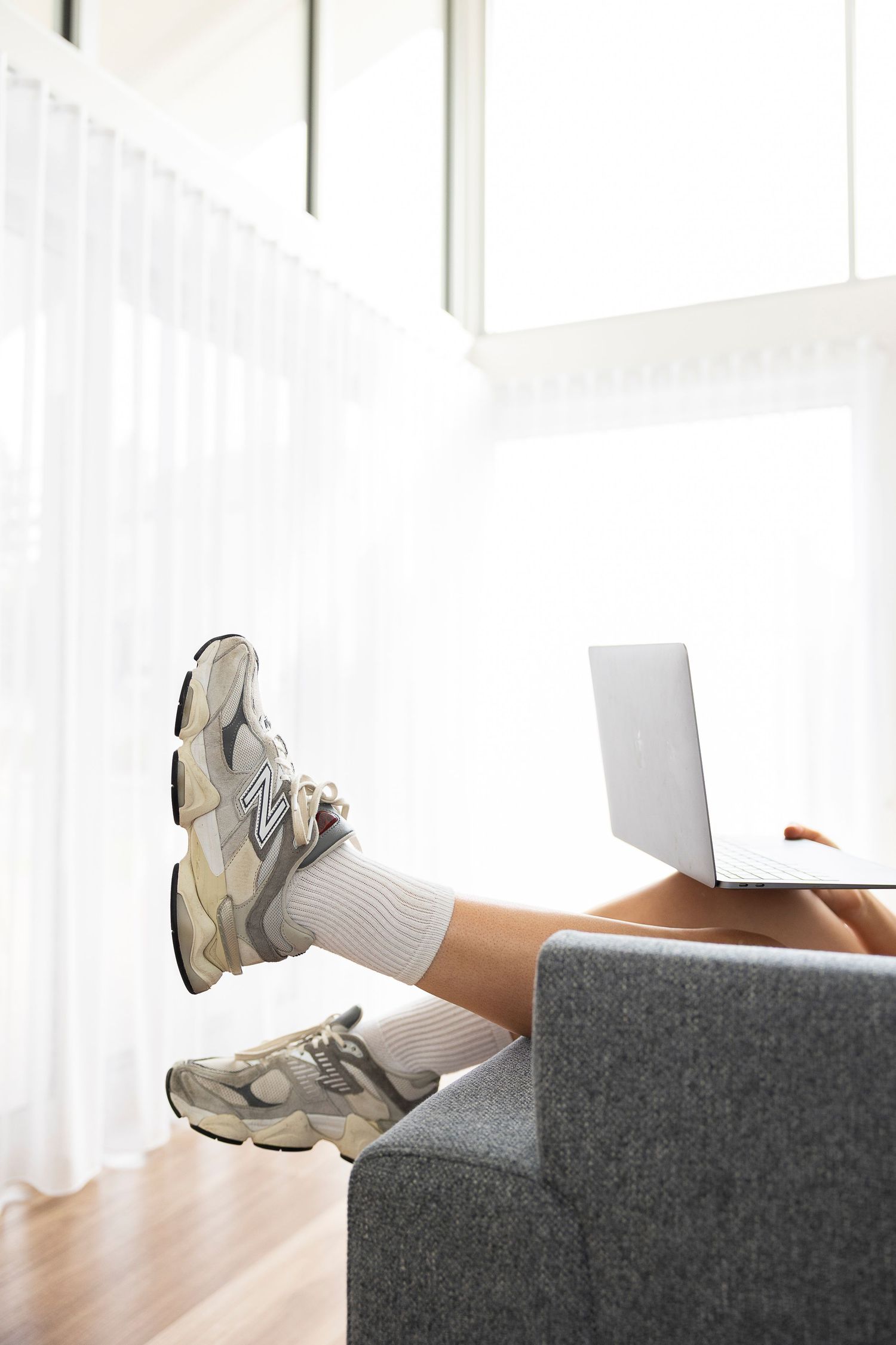 Person relaxing on gray sofa with legs up, wearing New Balance sneakers and white socks, using a laptop near sheer curtains.