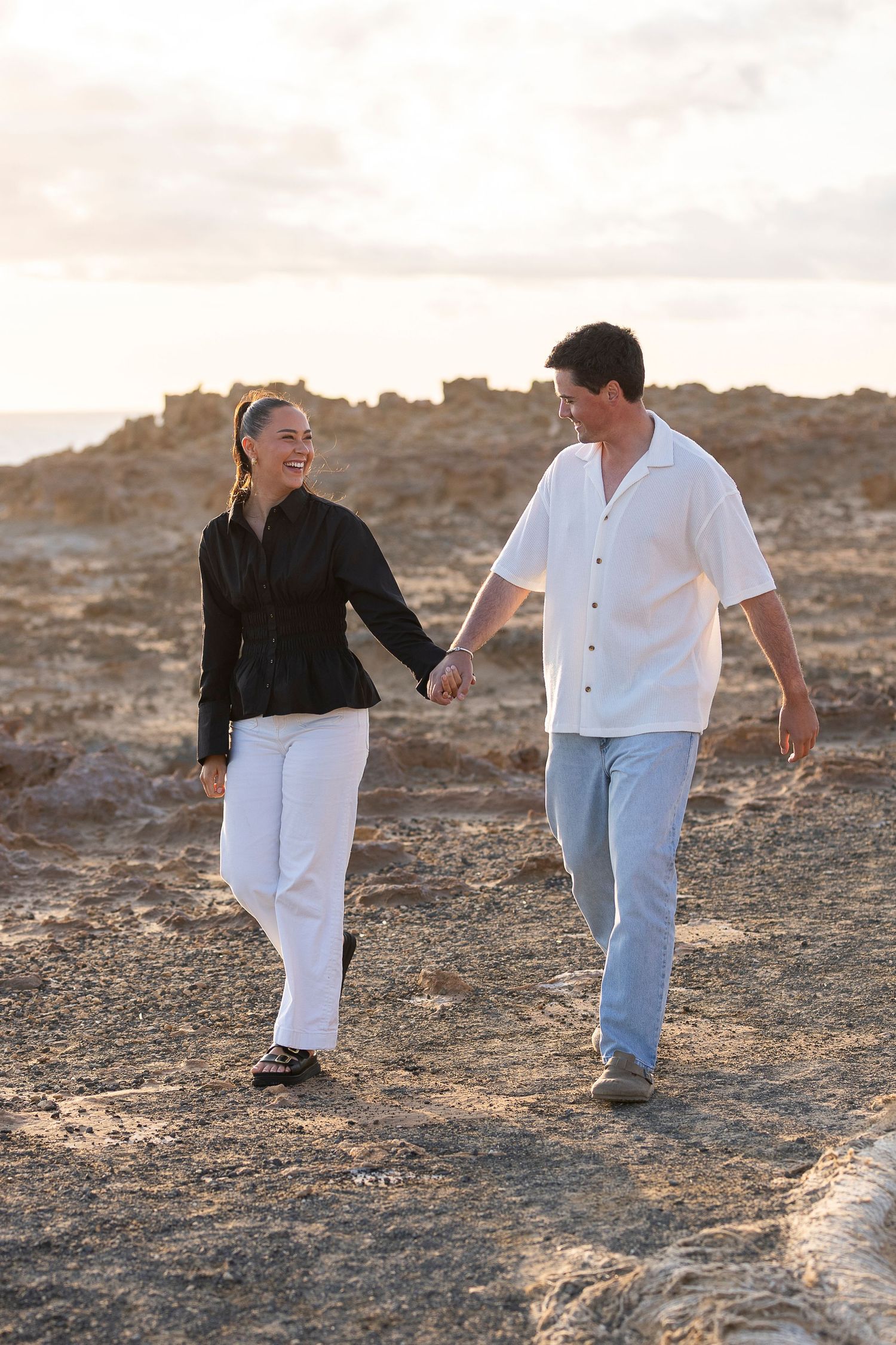 A playful couple walking and laughing together on a rocky coastal path at sunset.