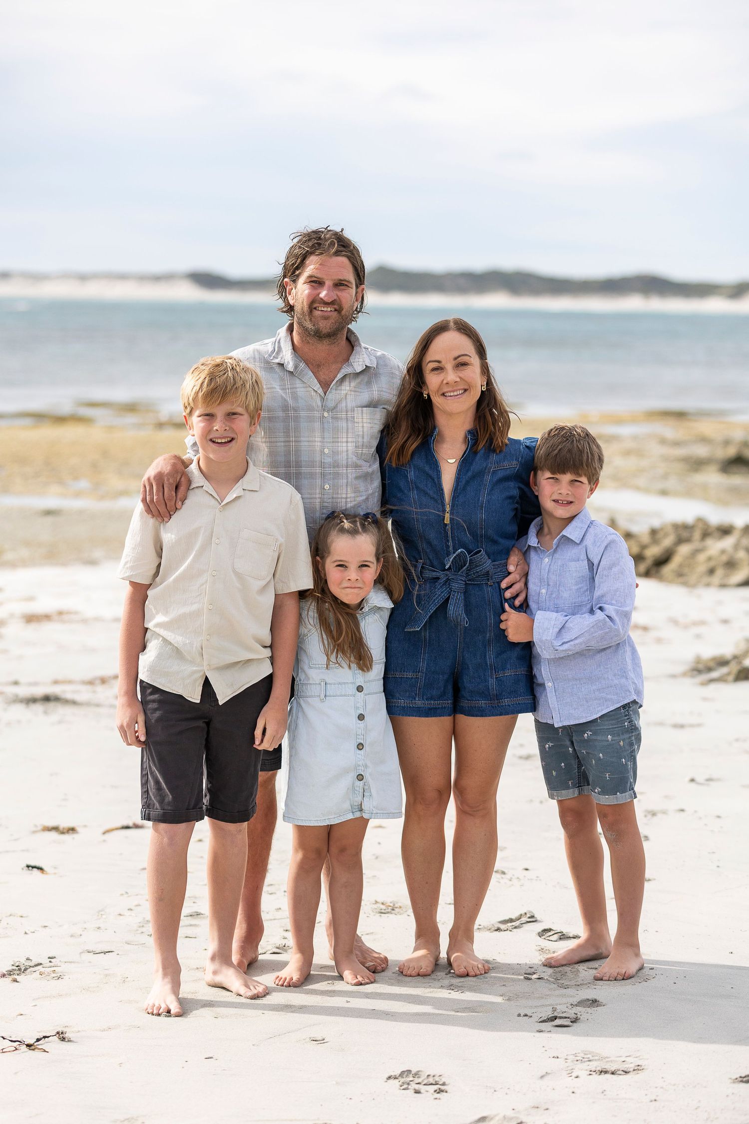 A family of five poses together barefoot on a sunny beach with ocean waves in the background.