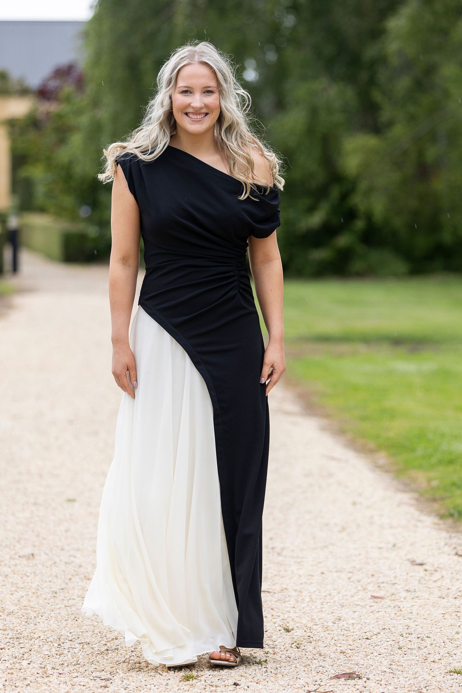 A person wearing a black and white contrast dress poses on a gravel path surrounded by greenery.