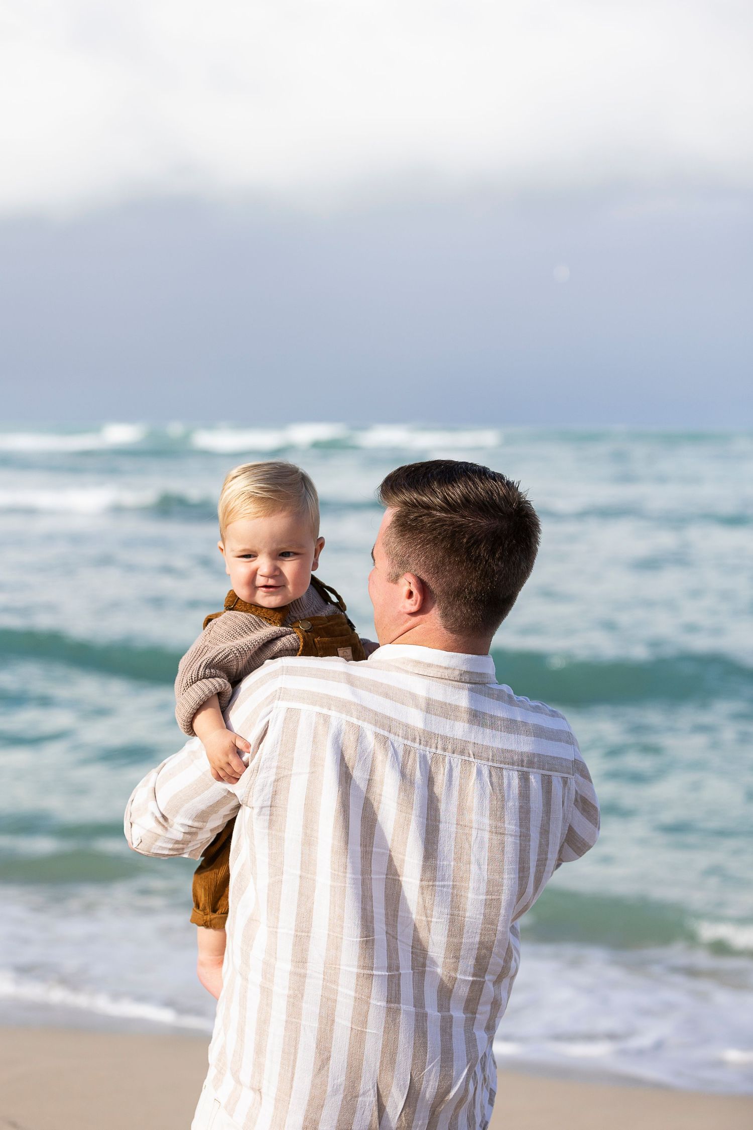 A tender beach moment with a baby in brown overalls being held against the ocean waves.