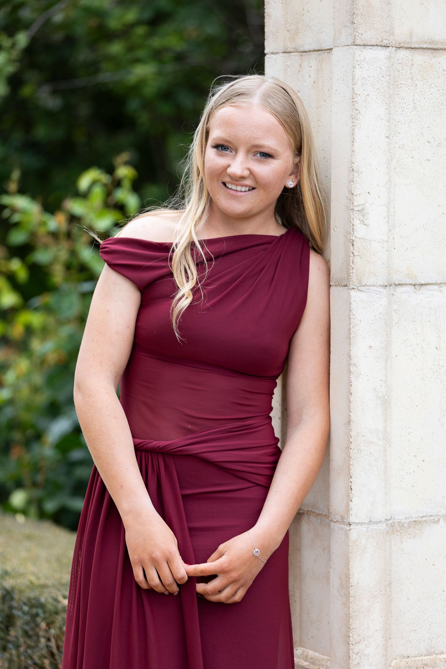 A person in burgundy dress poses against a light stone wall outdoors.