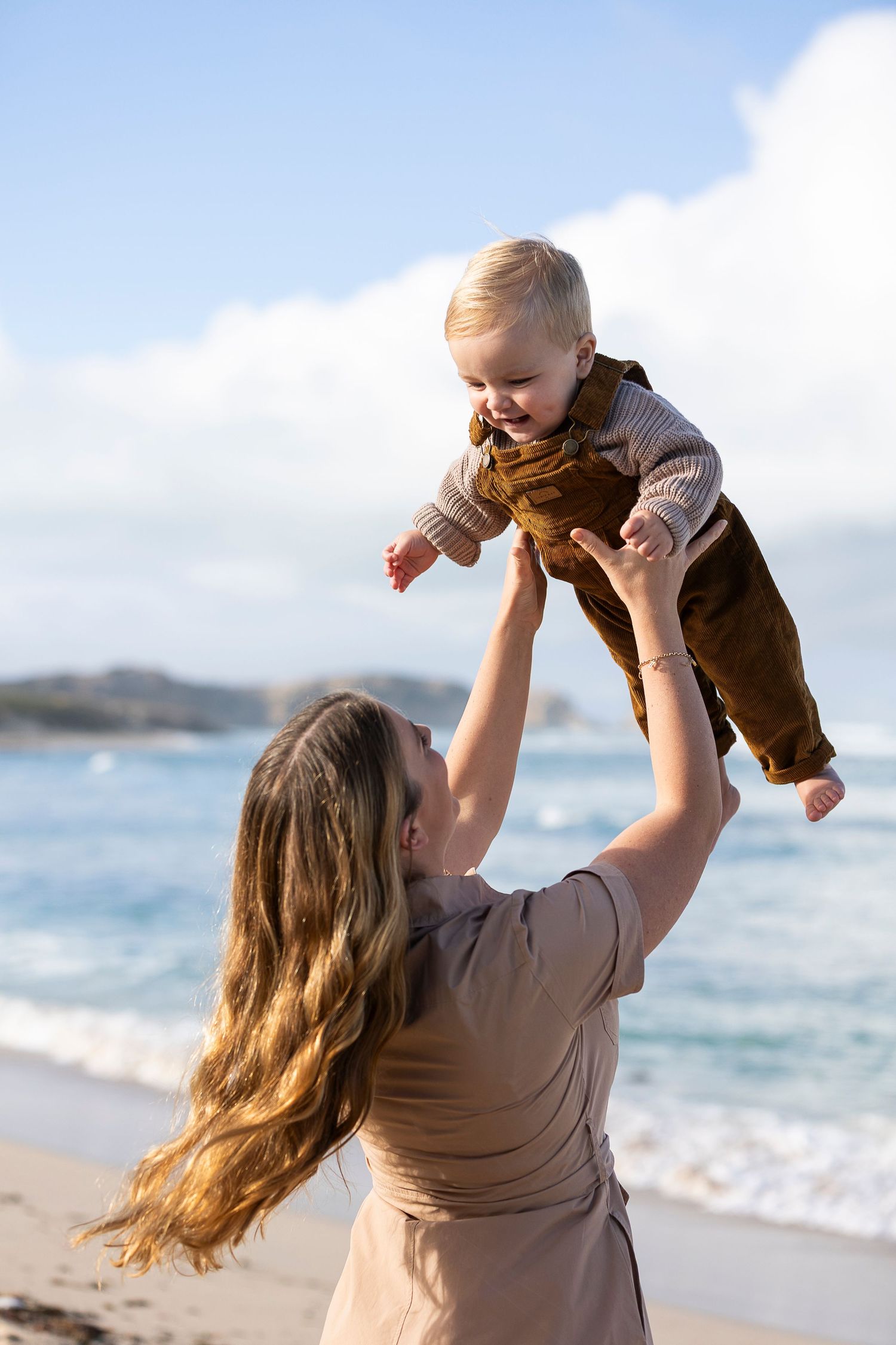 A person lifts a happy baby high in the air on a sunny beach.