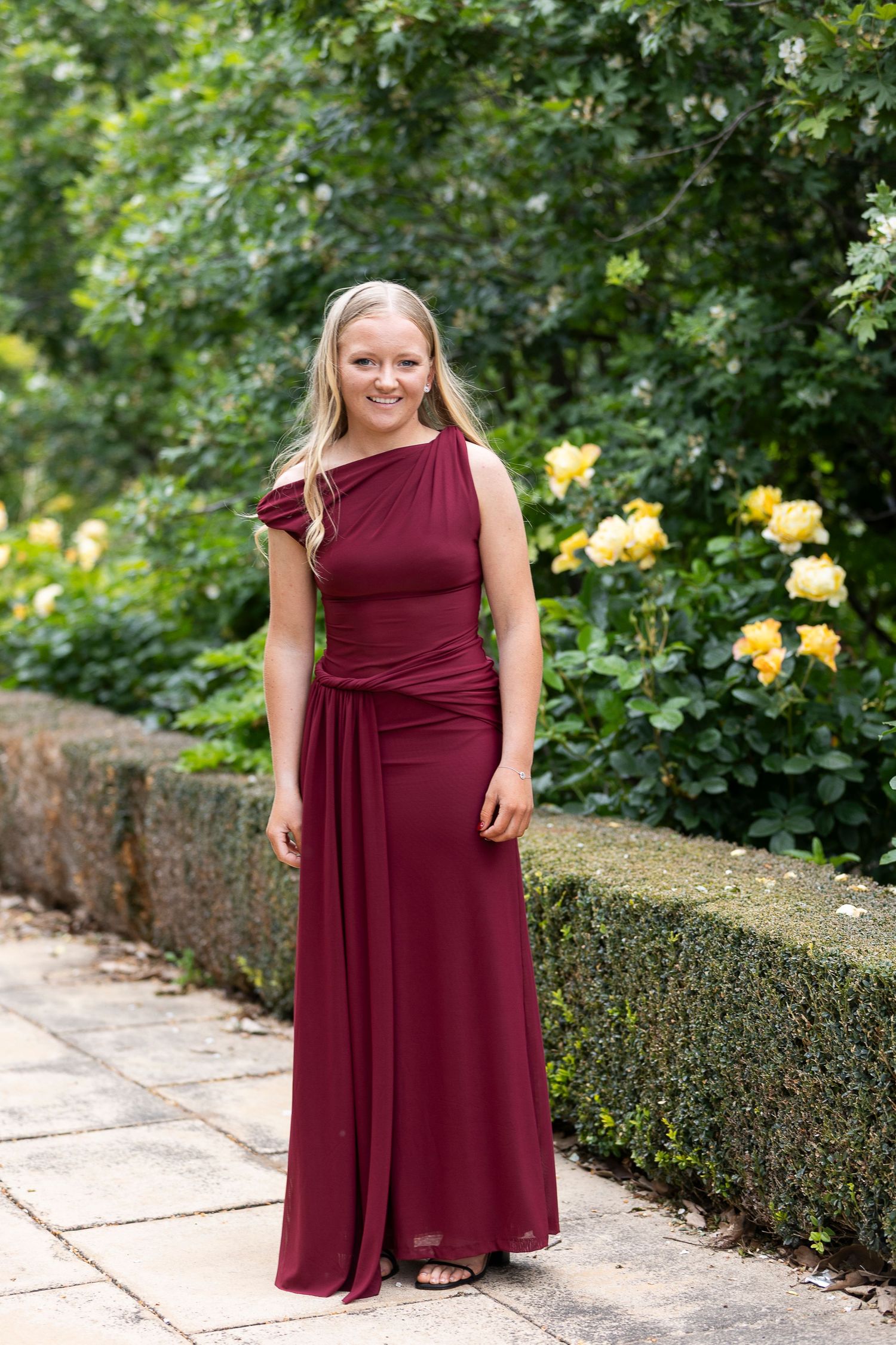 Burgundy formal dress shown in garden setting with yellow roses and manicured hedges.