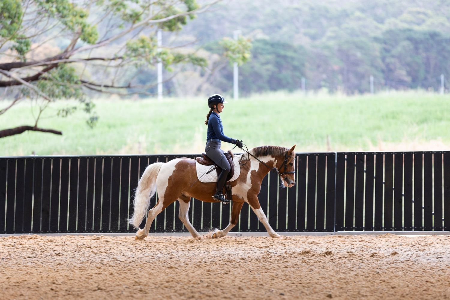 Horses and riders training in sequence along the rail of an indoor arena.