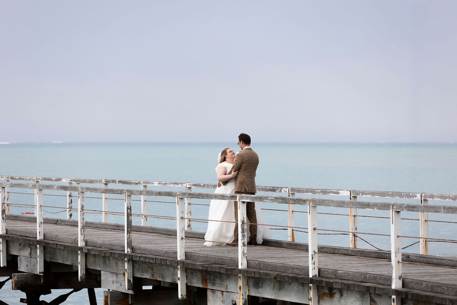 A couple stands together on a long wooden pier overlooking calm waters.