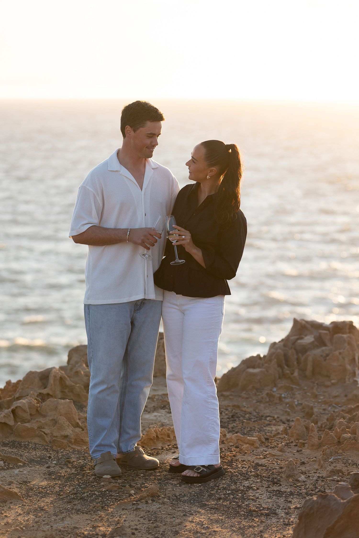 Couple sharing drinks on rocky beach at sunset.