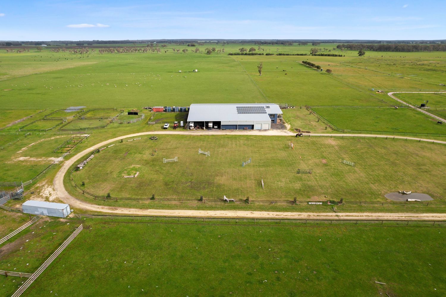 Aerial view of a rural farm property with a large steel shed surrounded by lush green paddocks and a winding gravel driveway.