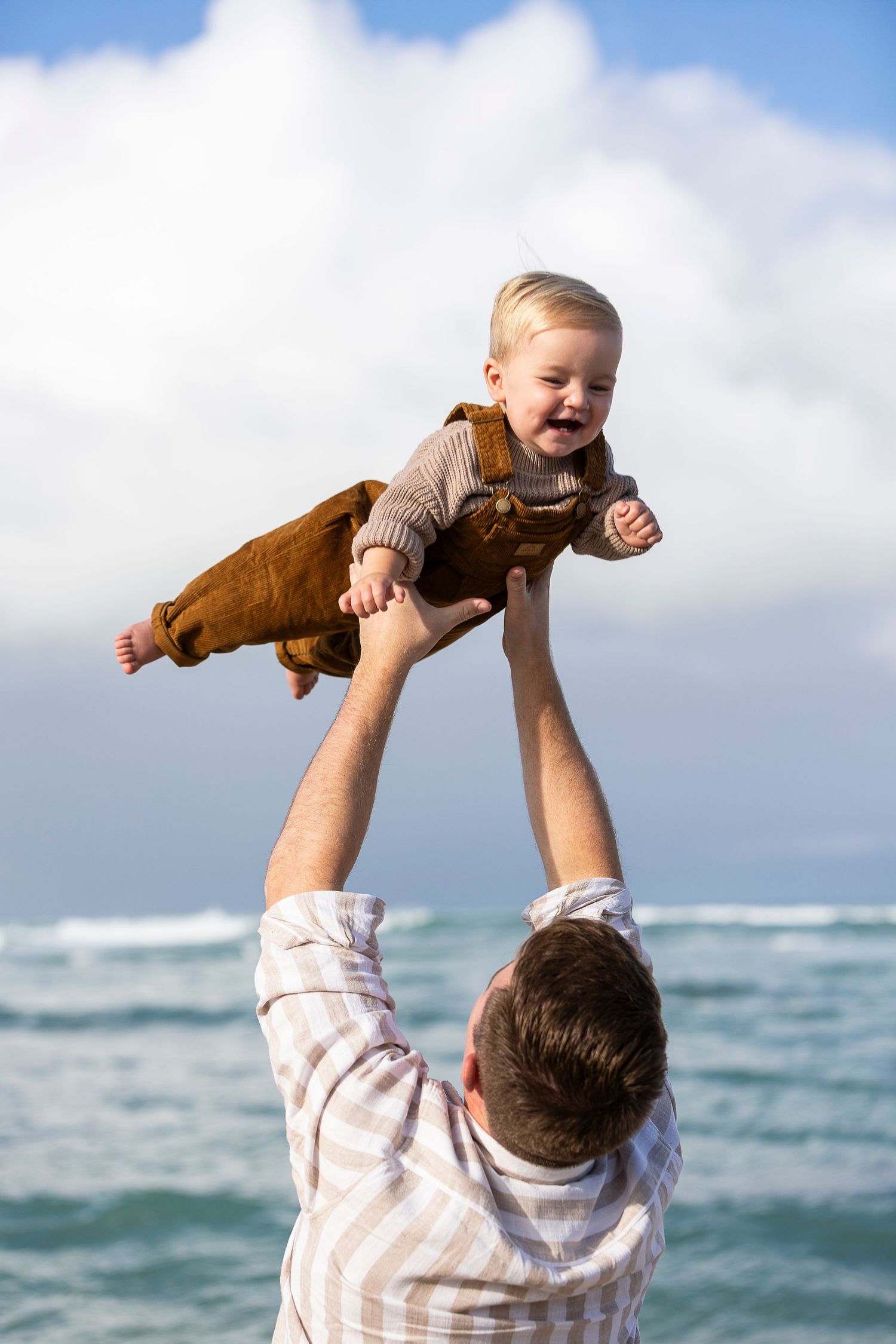 A joyful moment at the beach as a baby in brown overalls is lifted high against a cloudy ocean backdrop.