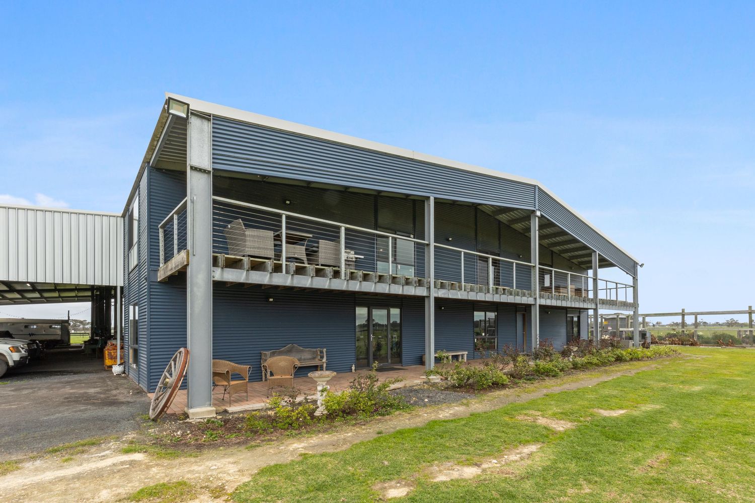 Two-story blue steel farm building with wraparound balcony, large windows, and green lawn under clear blue sky.