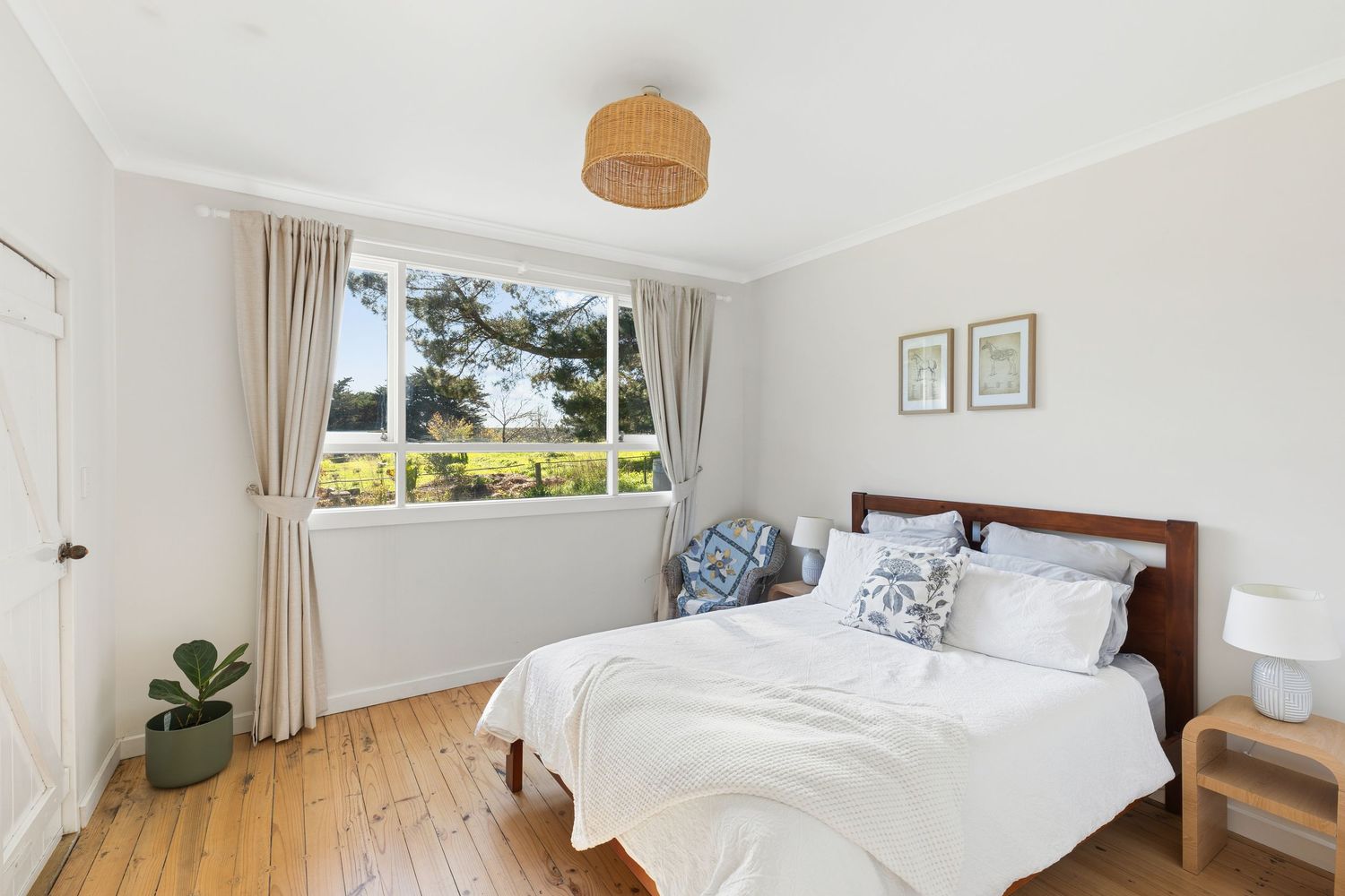 Bright bedroom with hardwood floors, white bedding, timber bed frame, rattan pendant light, and a window with garden views.
