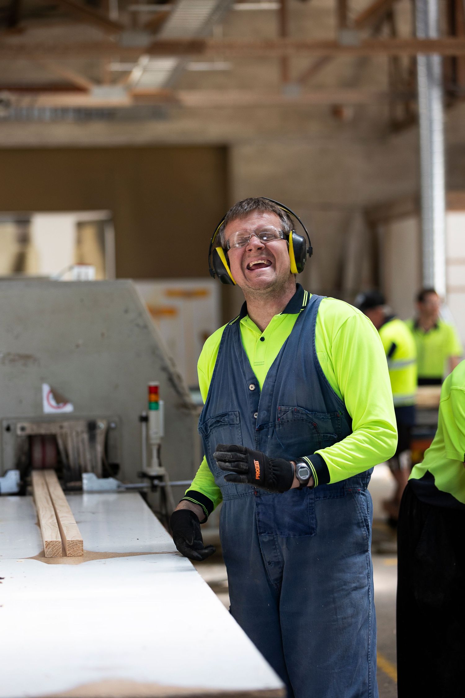 Factory workers in safety vests and hearing protection work with wood materials in an industrial setting.