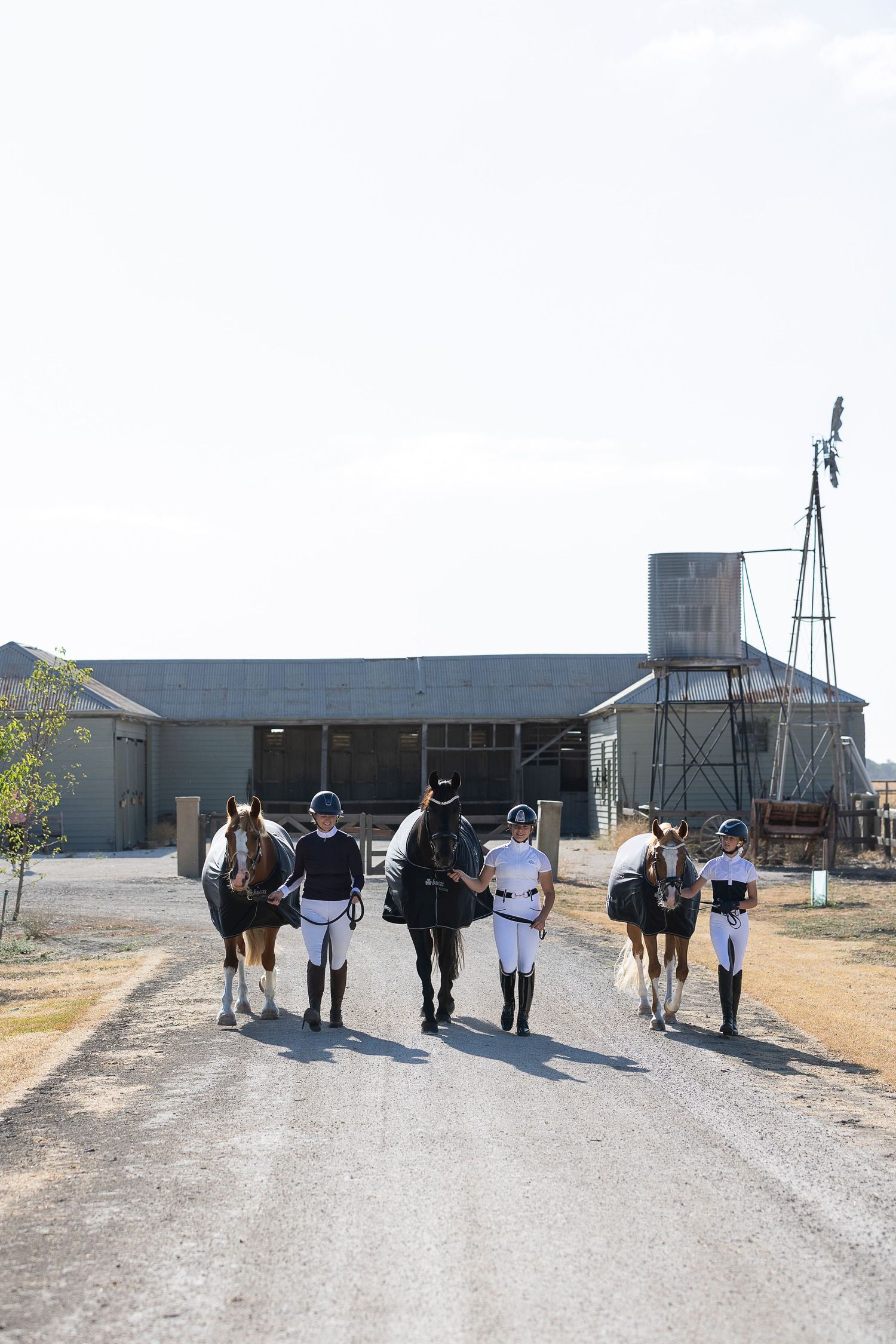 Group of equestrians walking with their horses on a path near farm buildings.