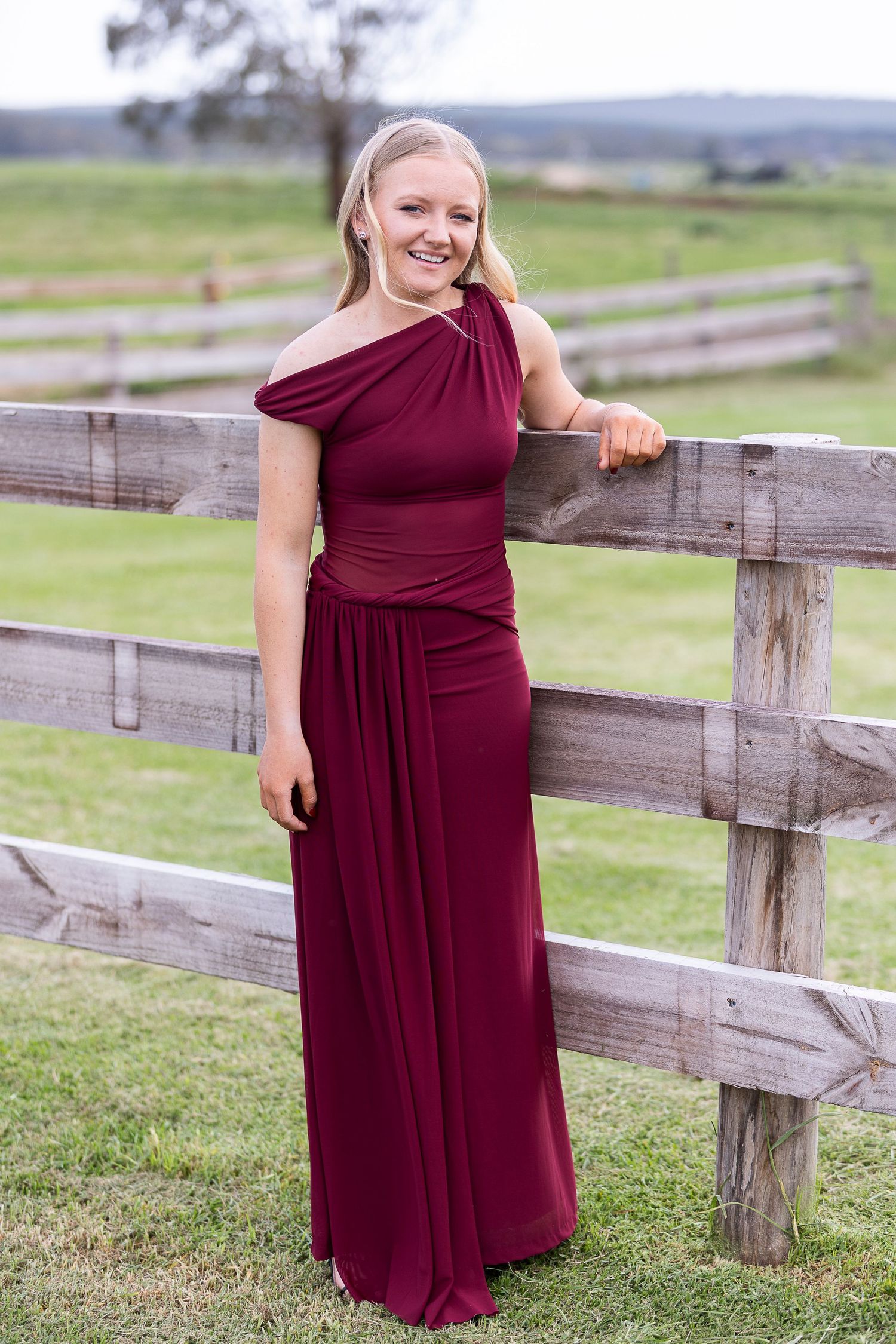 A woman in a burgundy evening gown poses by a wooden fence in a rural setting.