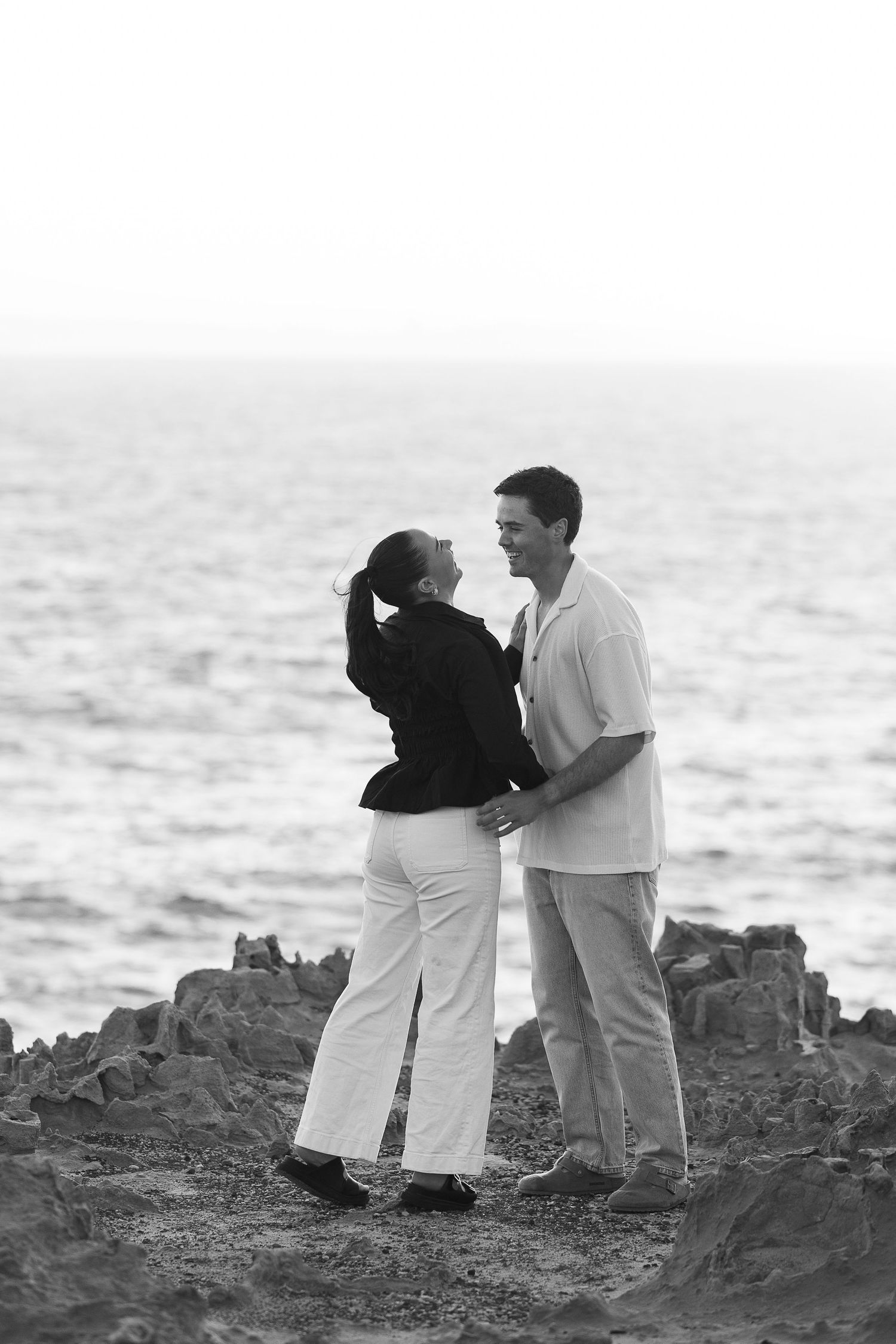 Couple stands together on rocky coastal outcrop at sunset.