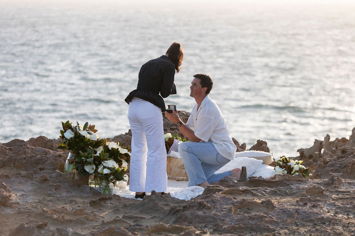 A romantic sunset proposal moment captured on rocky coastal cliffs overlooking the ocean.