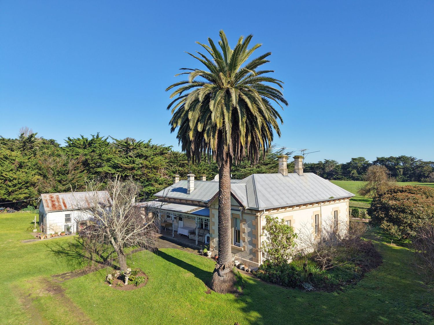 Aerial view of a historic stone homestead with a tall palm tree, lush green lawn, and surrounding trees under a clear blue sky.
