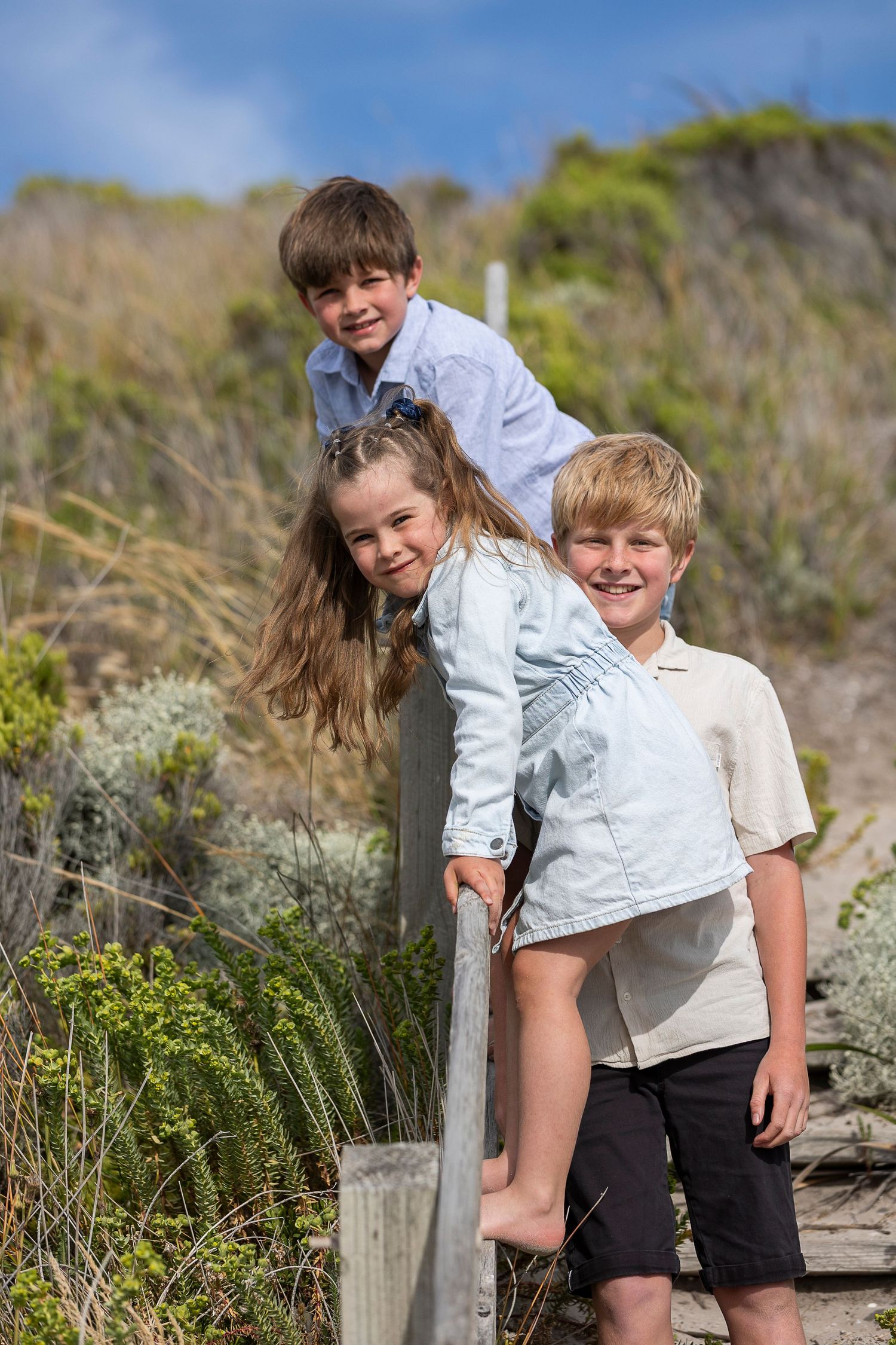 Three children play and pose together on coastal hiking trail with vegetation.