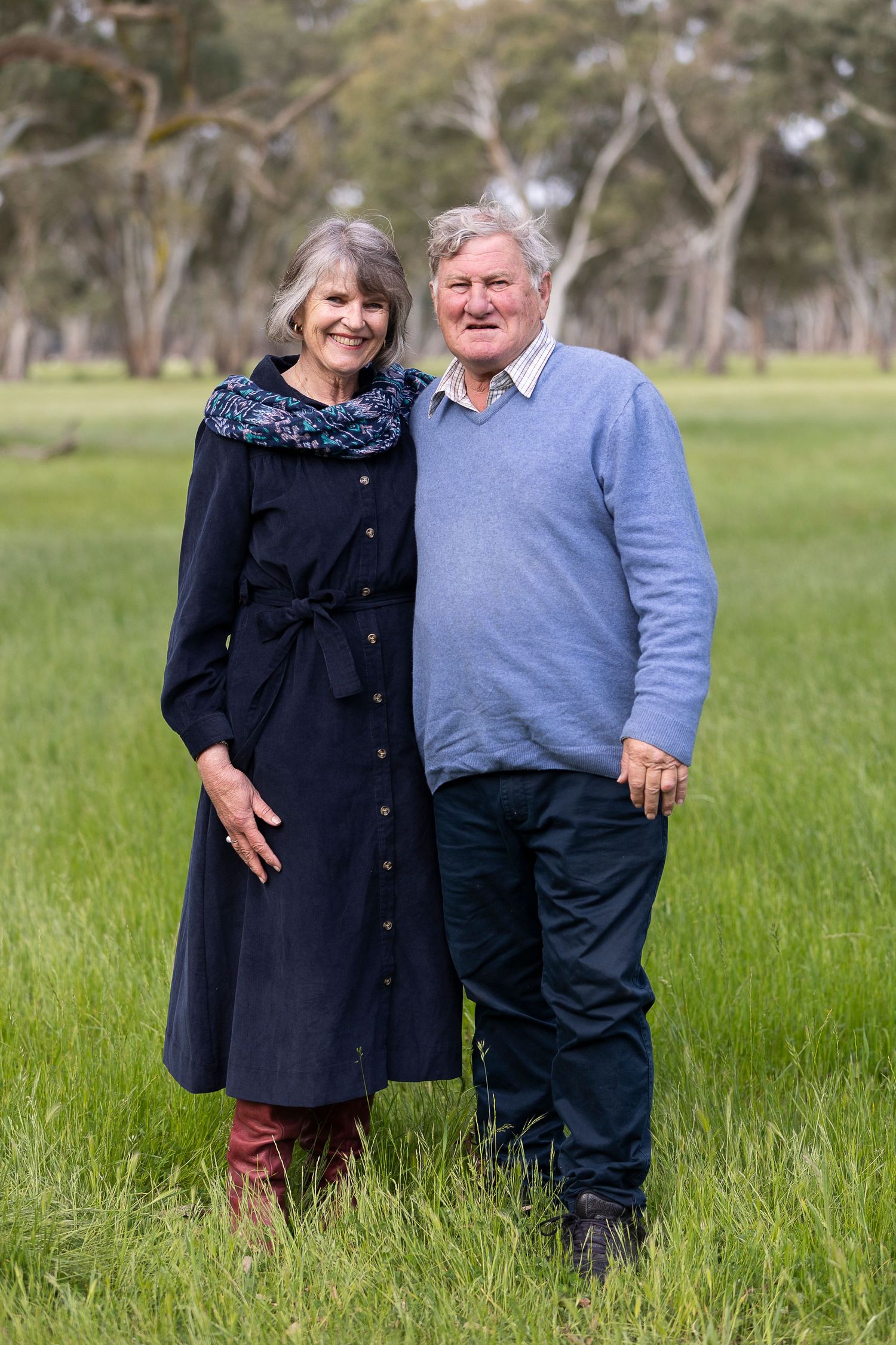 A couple stands together in a grassy field wearing navy and grey casual clothing.