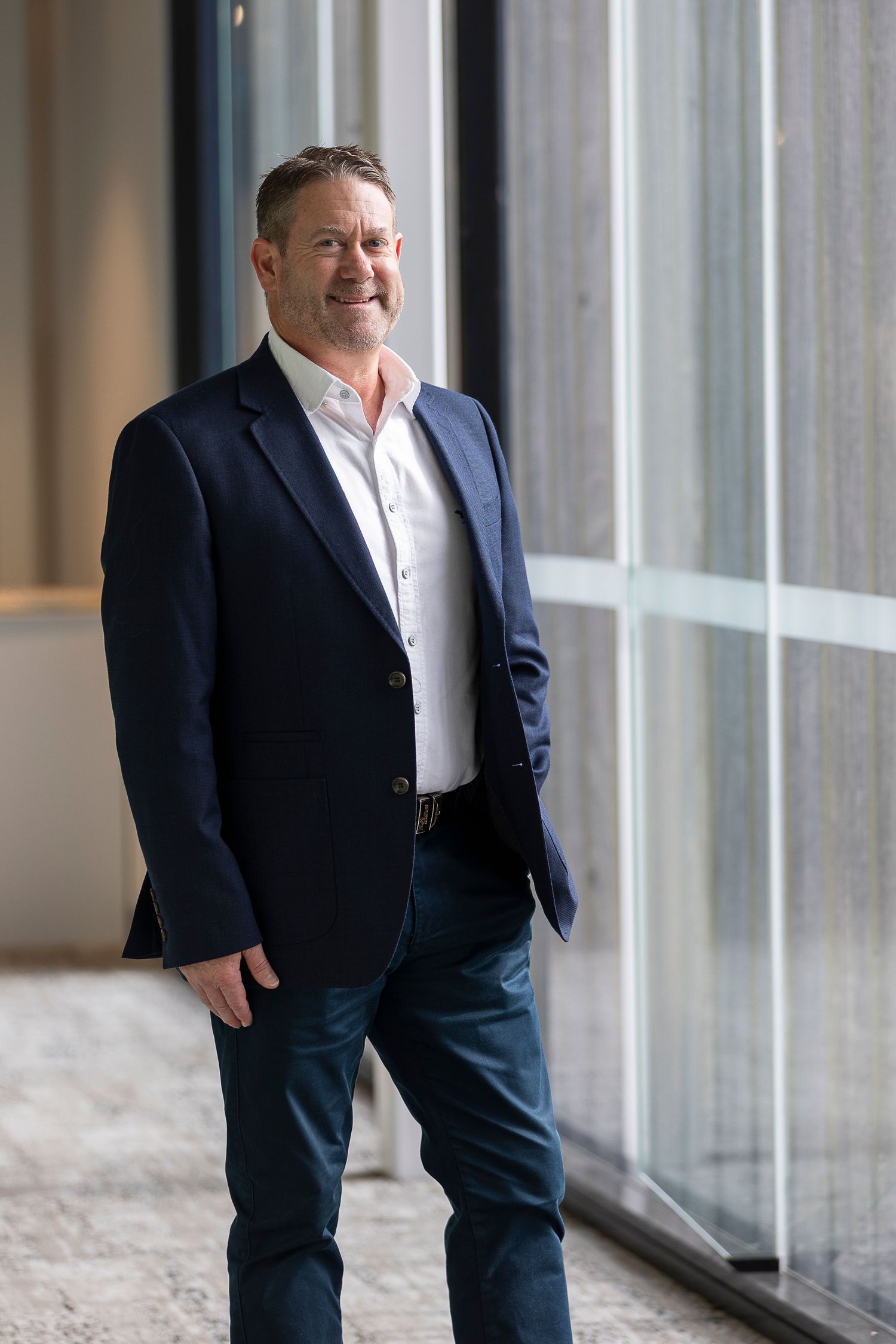 Professional man in navy blazer and jeans smiling confidently near large windows in modern office building.