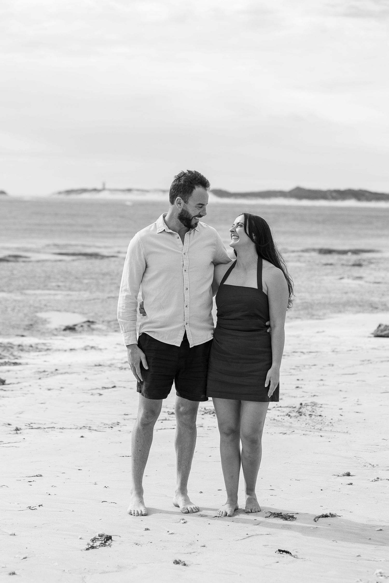 A couple stands together barefoot on a beach in black and white.