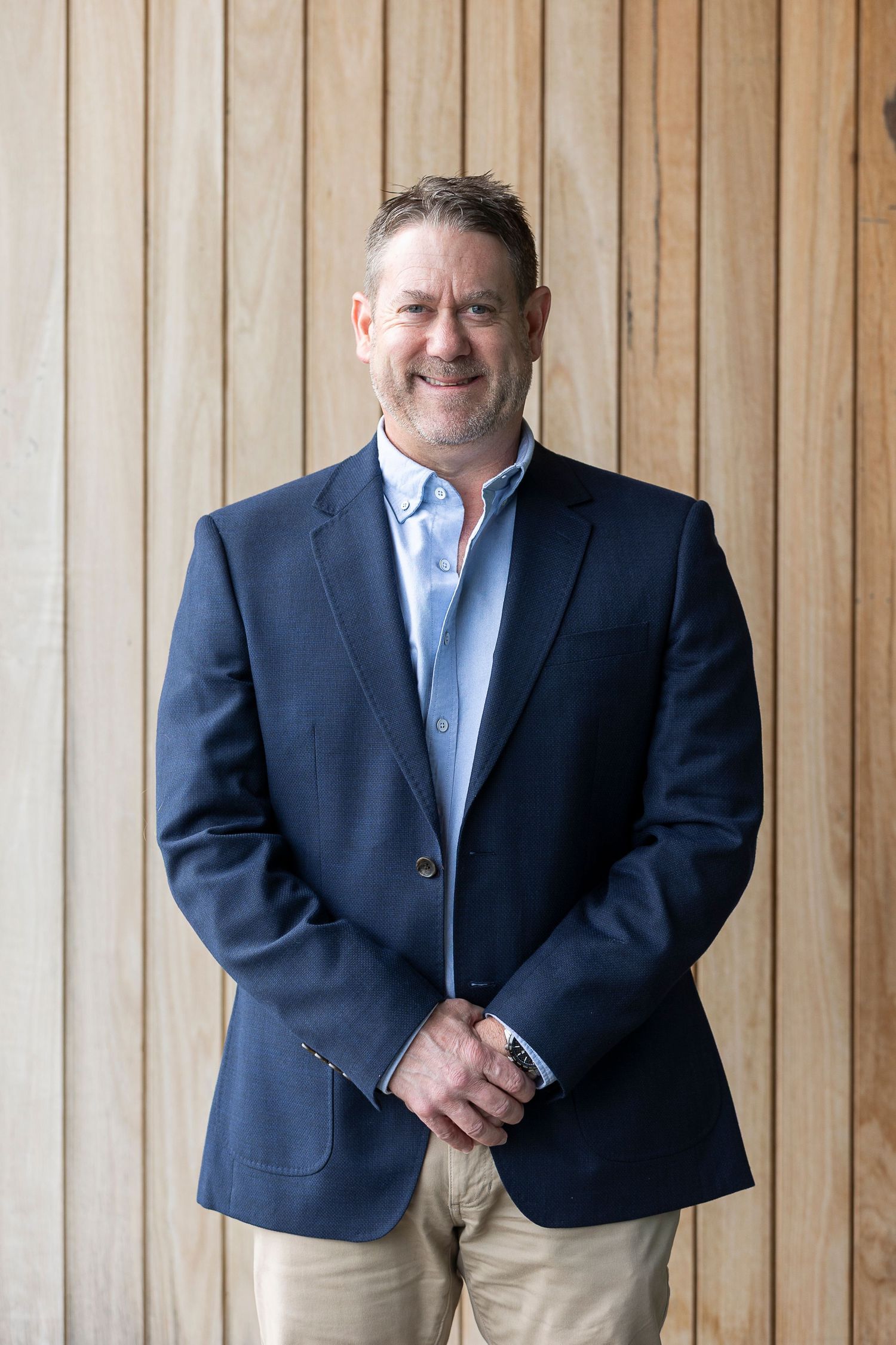 Smiling middle-aged man in navy blazer and light blue shirt standing against wooden wall background.