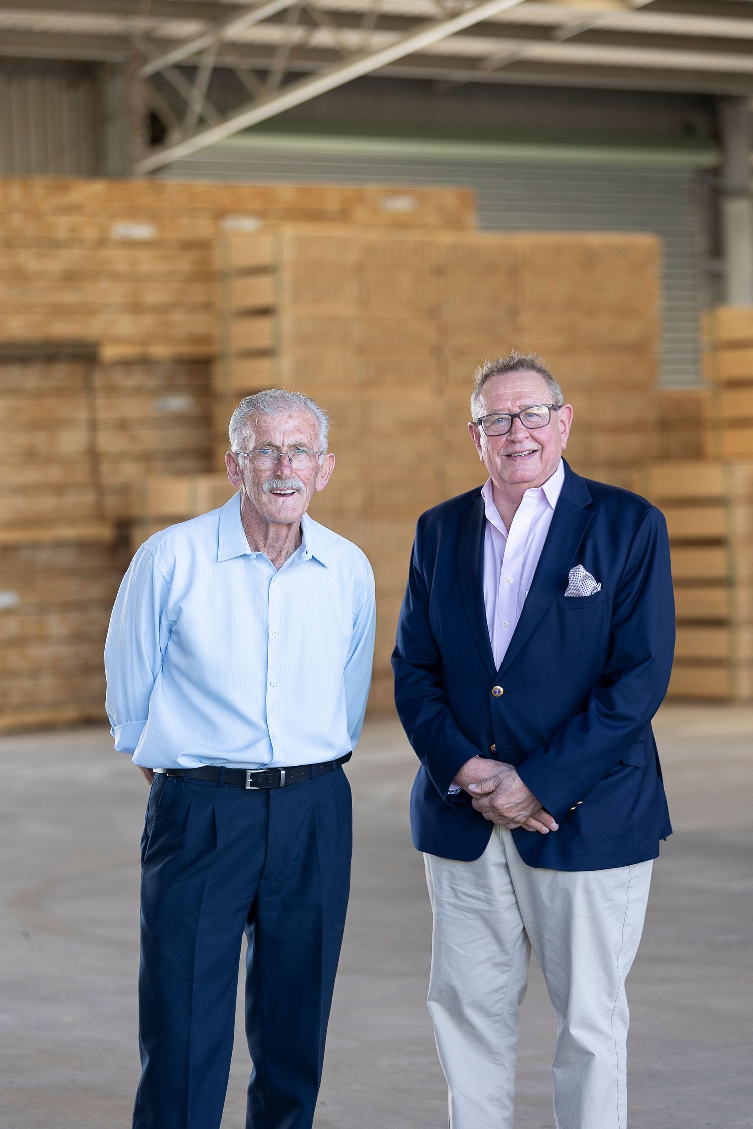 Two businessmen standing together in a warehouse with stacked lumber in the background.