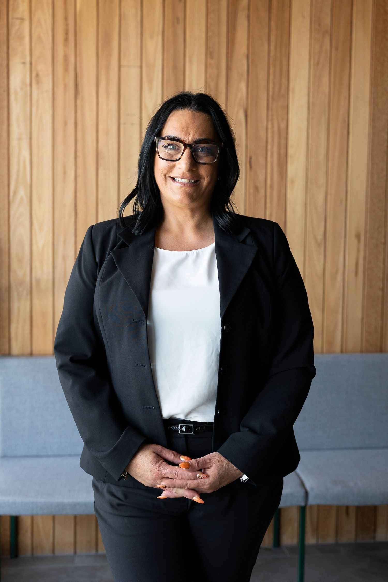 Professional in business attire seated against wooden paneled wall.