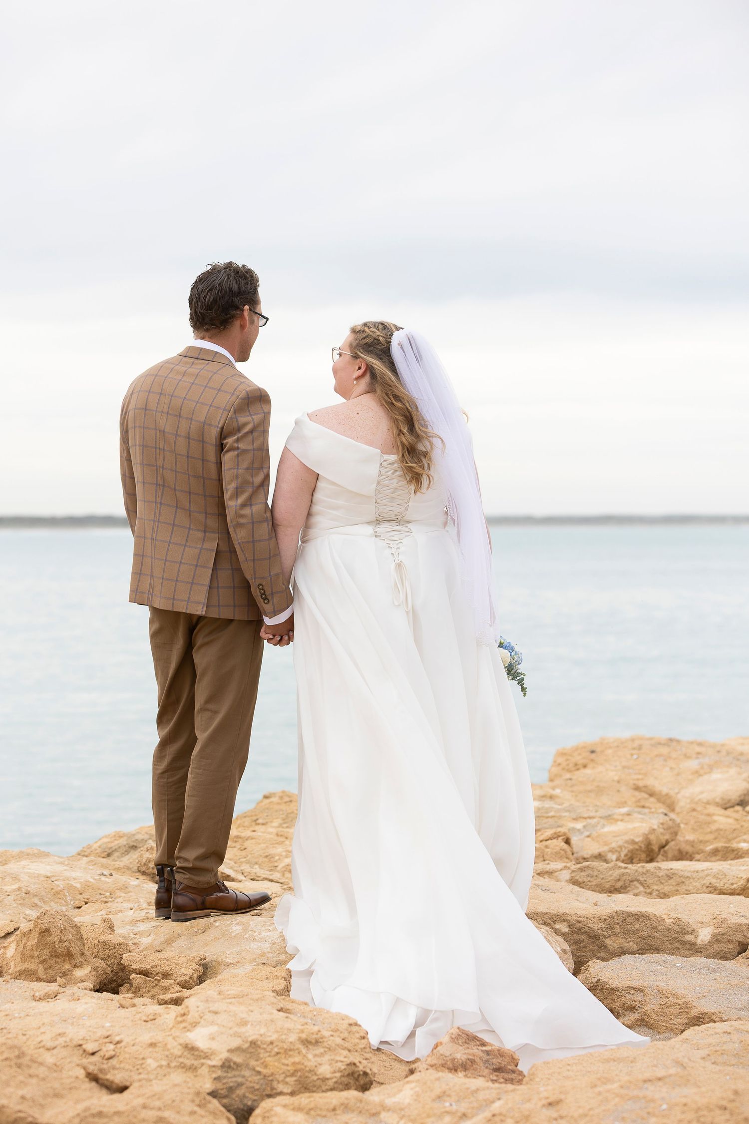 A couple in wedding attire stands together on rocky coastal cliffs overlooking the ocean.