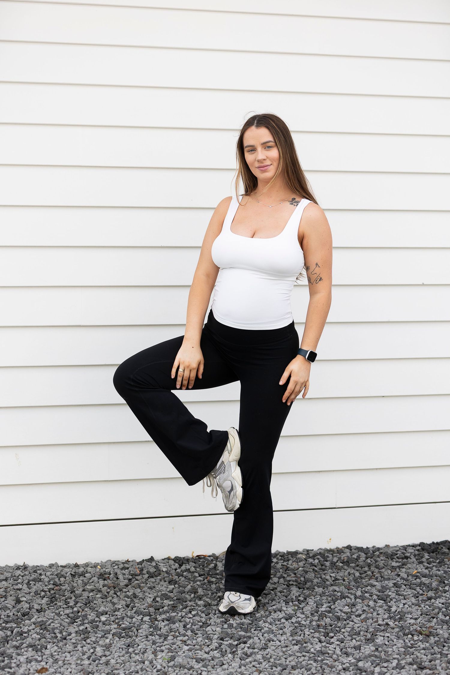 Pregnant woman in white tank top and black flare pants leaning against white wall outdoors.