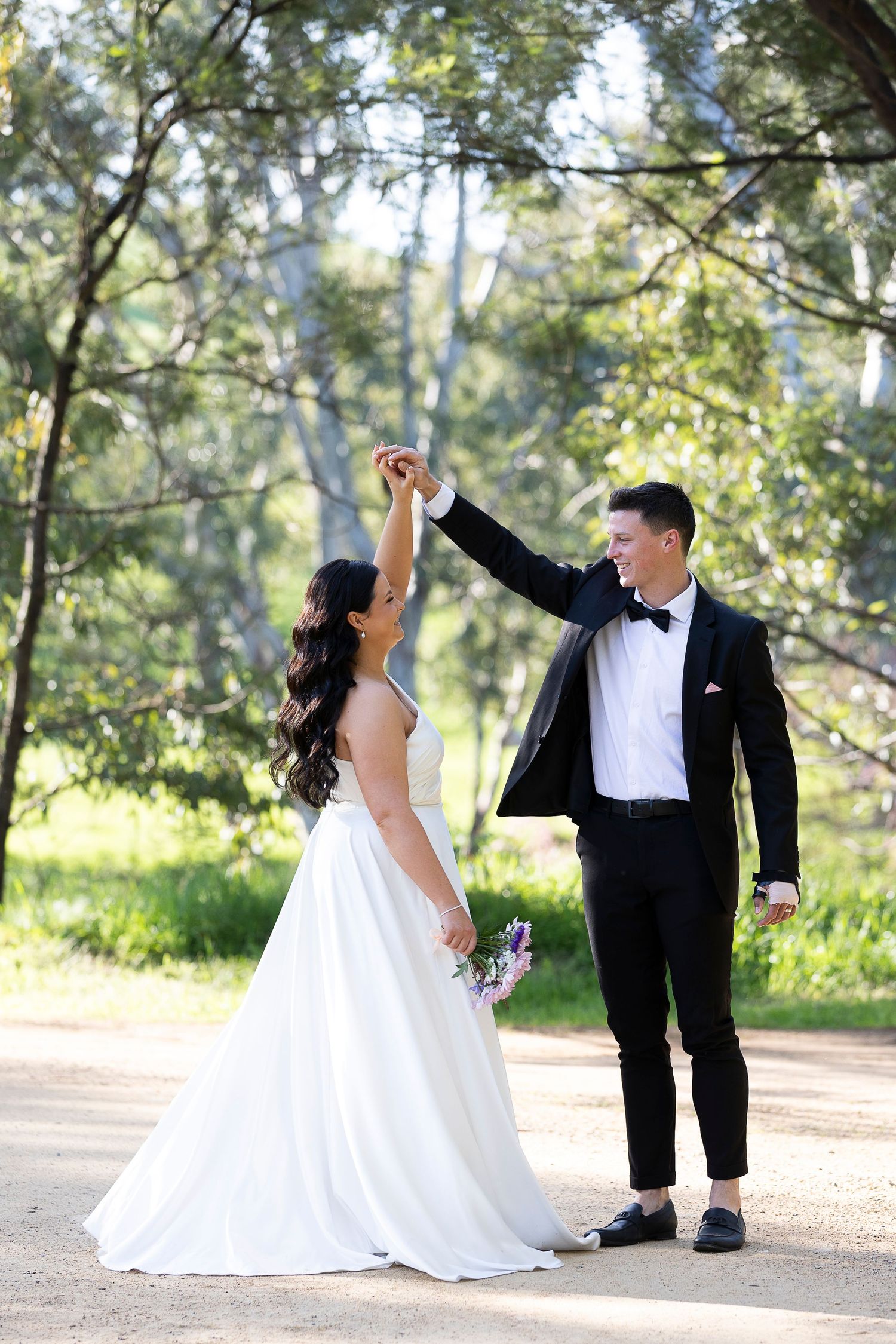 Playful wedding couple dances and twirls on a tree-lined path.