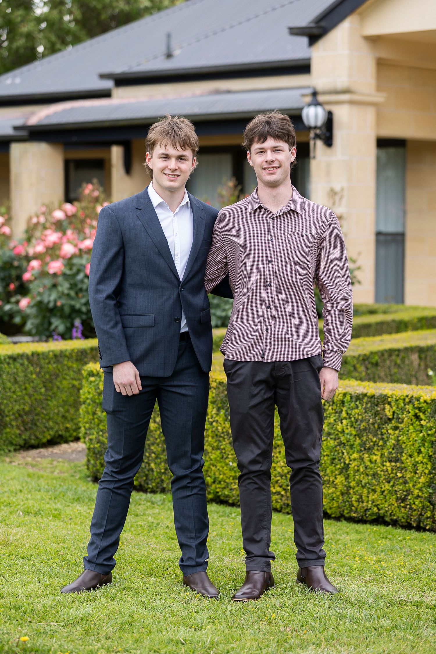 Two young men in suits stand together on a lawn with a house and garden in the background.