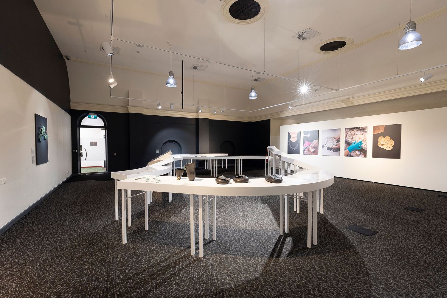 Wide-angle view of an art gallery space with minimalist white display tables and spotlighting.
