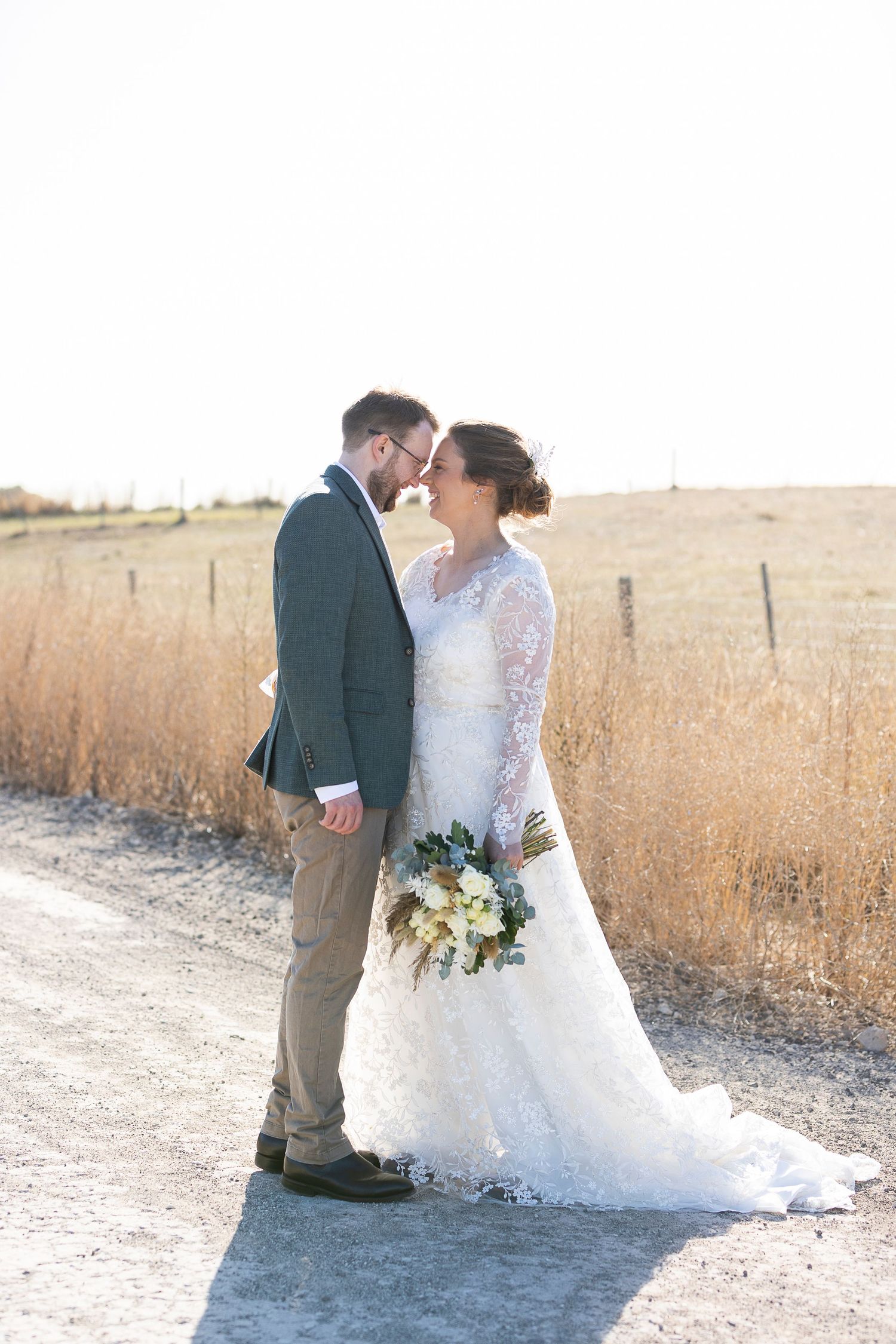 Wedding couple poses along a dirt path surrounded by golden autumn fields.