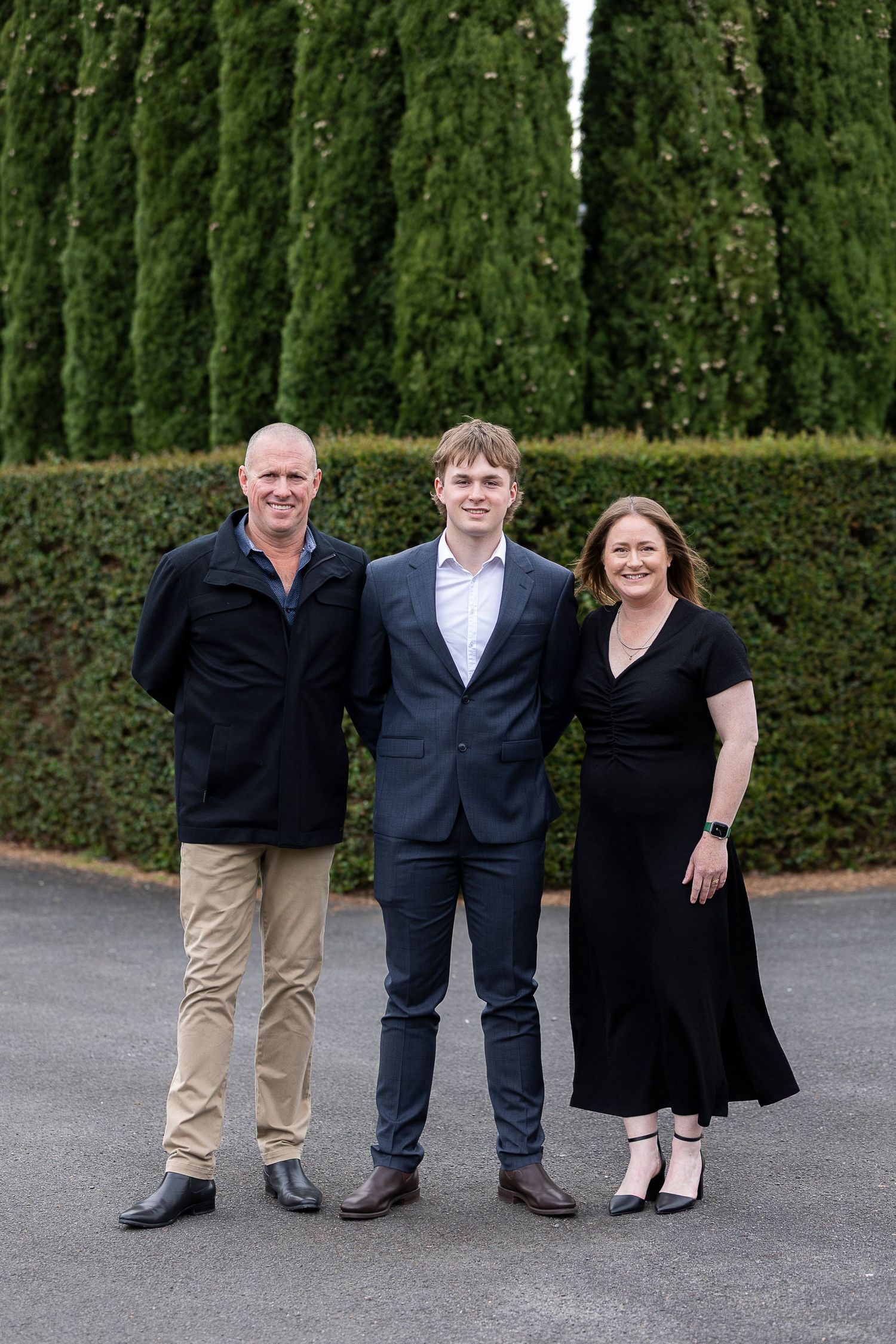 Three people in formal attire stand in front of a tall green hedge.