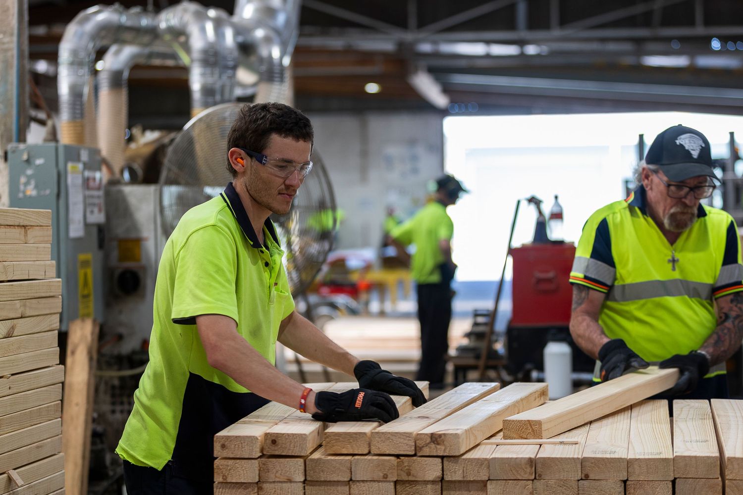 Workers in high-visibility gear assemble wooden components in a bright manufacturing facility.